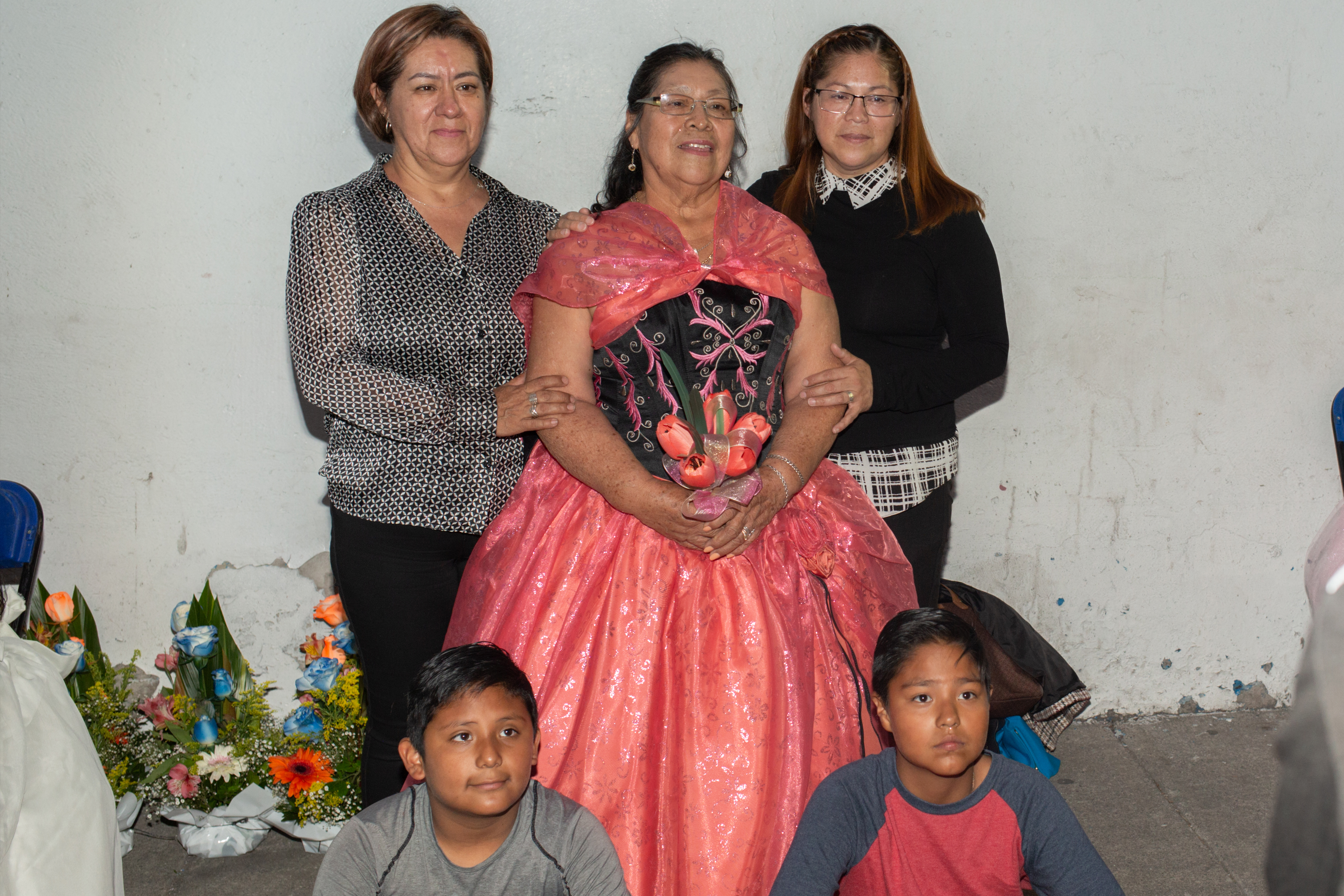 A woman in a hot pink ballgown with a blue bodice carries a bouquet of matching pink tulips as she is surrounded by her family, two women on either side of her and a pair of young boys at her feet. Together, they pose for a photo.