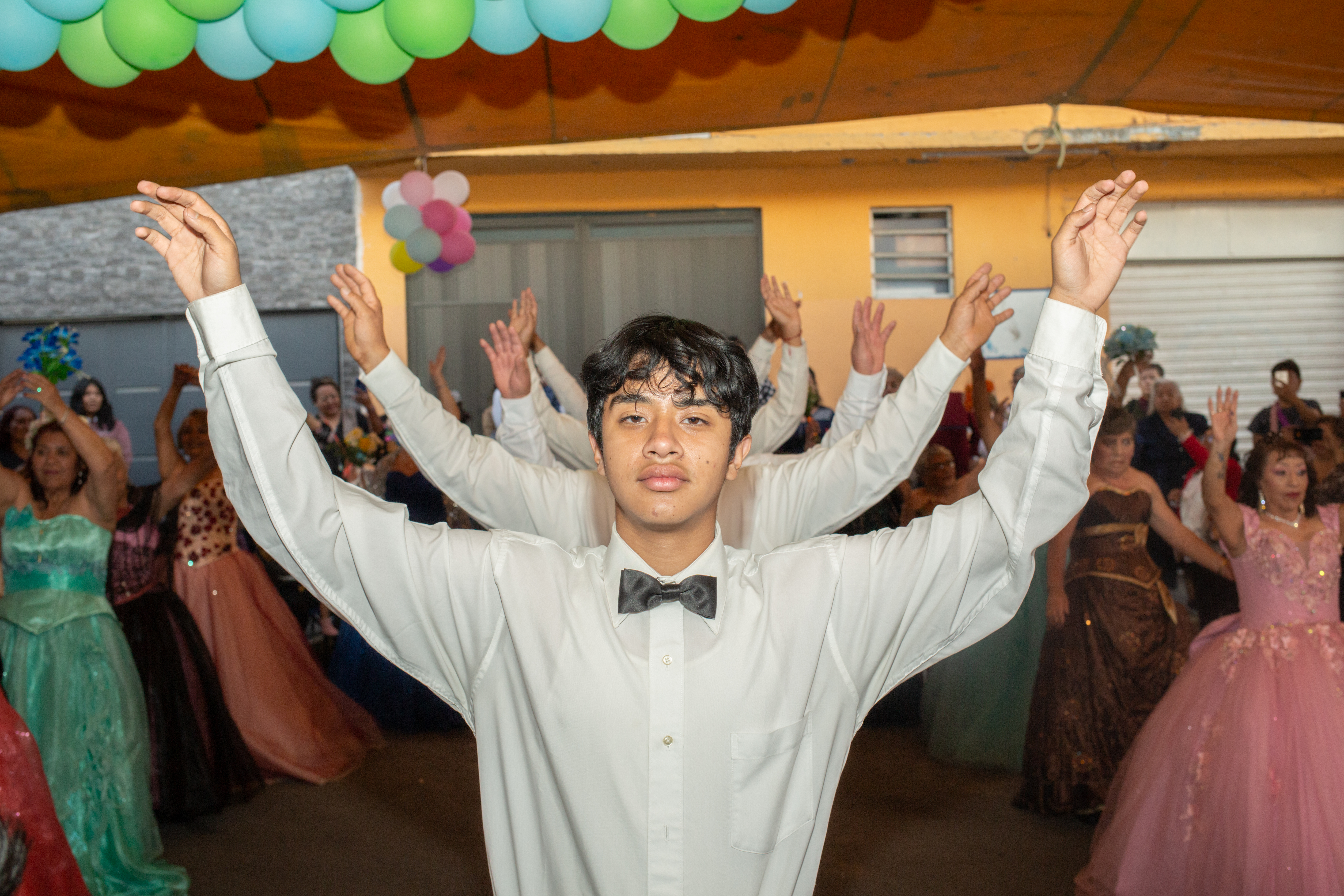 Young men, dressed in white dress shirts and black bowties, lift both of their arms in the air as they prepare to dance under a tent decorated with green and blue balloons.