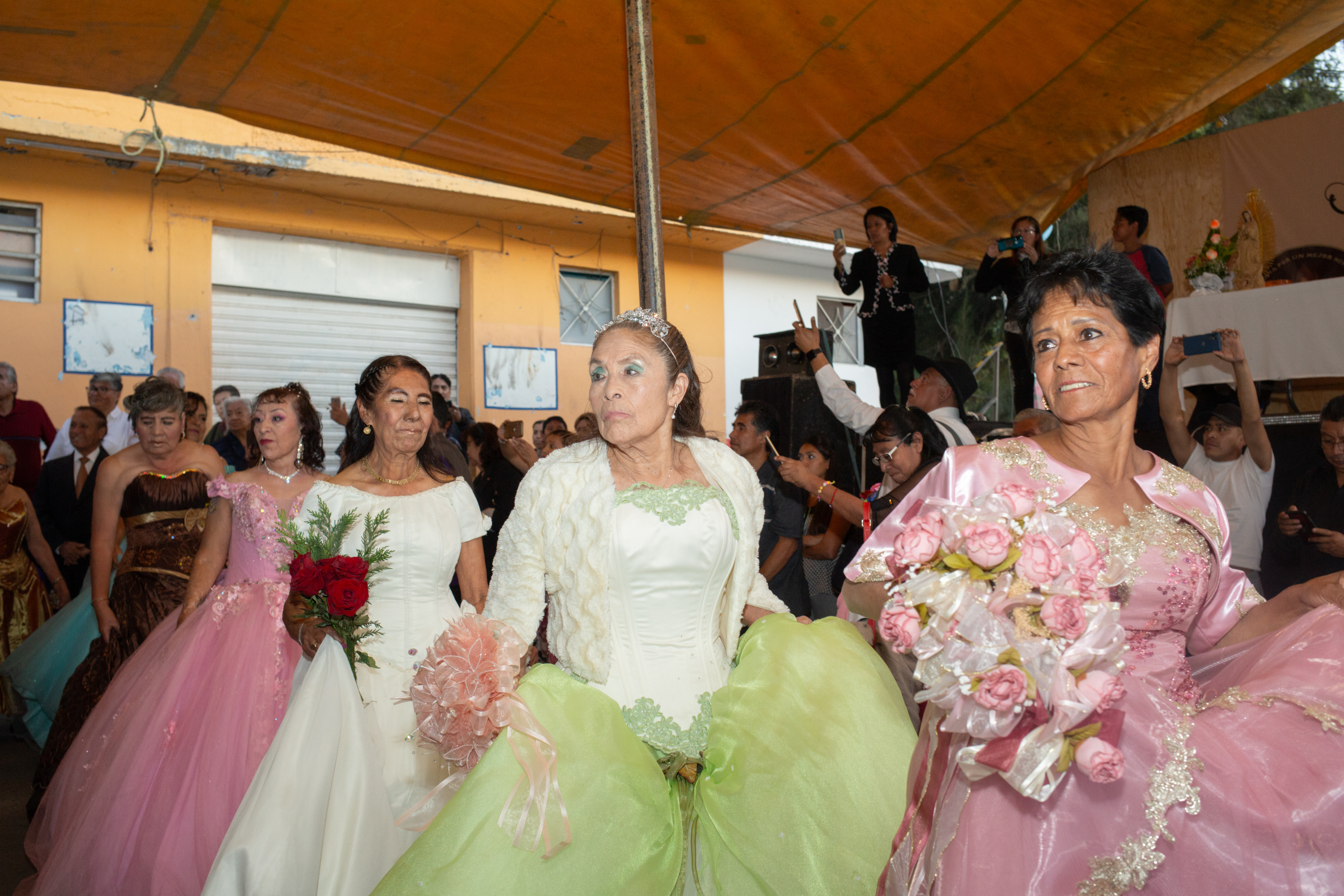 A line of women, dressed in poofy ballgowns of pink, white and pastel green, carry bouquets and assemble in a line, carrying their skirts in their free hand as they prepare to dance under the cover of an outdoor tent.