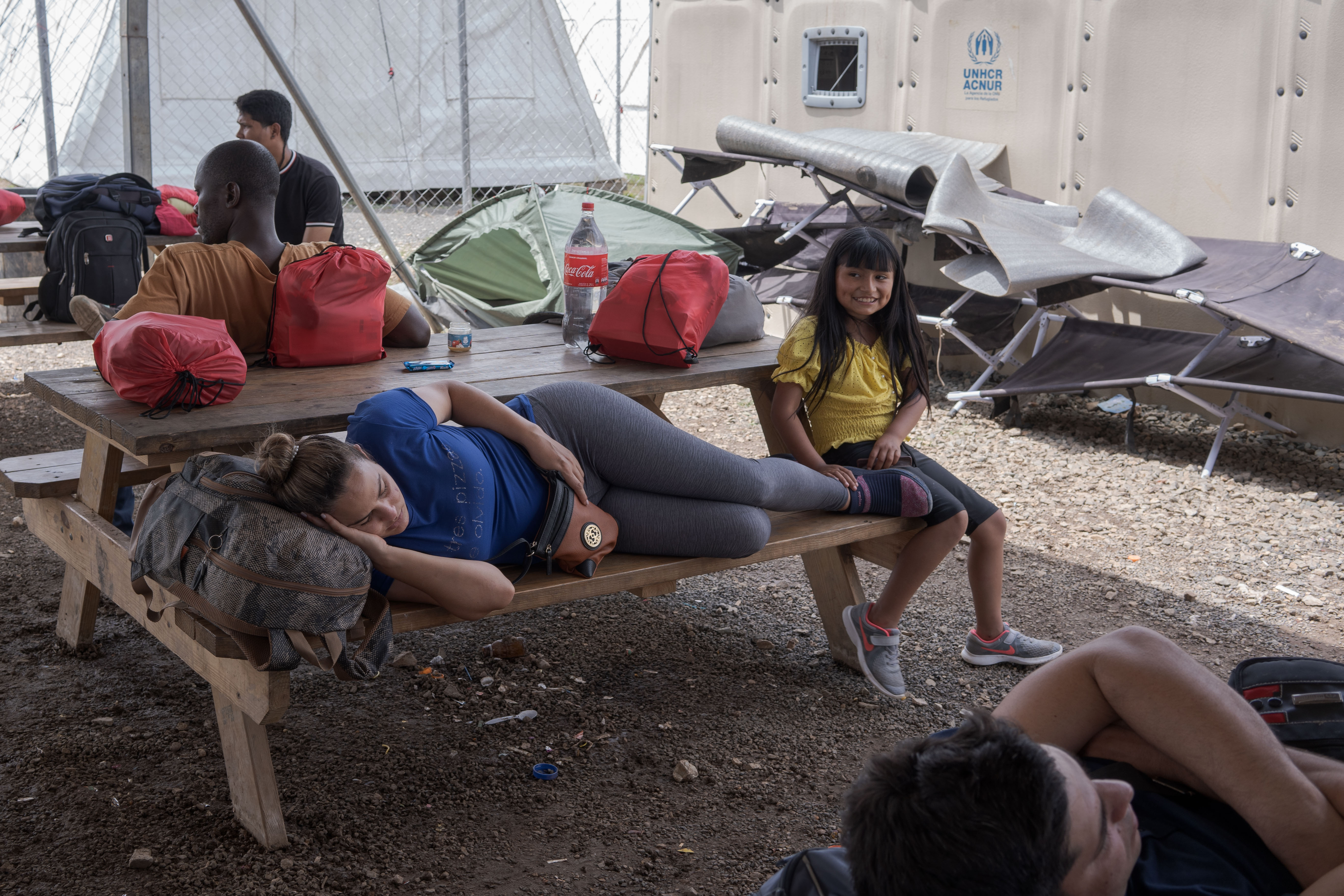 A woman lies down on an outdoor cot, while a child sits perched her feet, smiling at the camera.