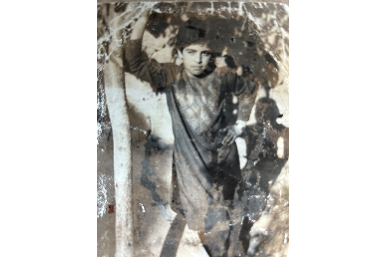 An old photograph of a boy standing with one arm on a thin tree trunk.