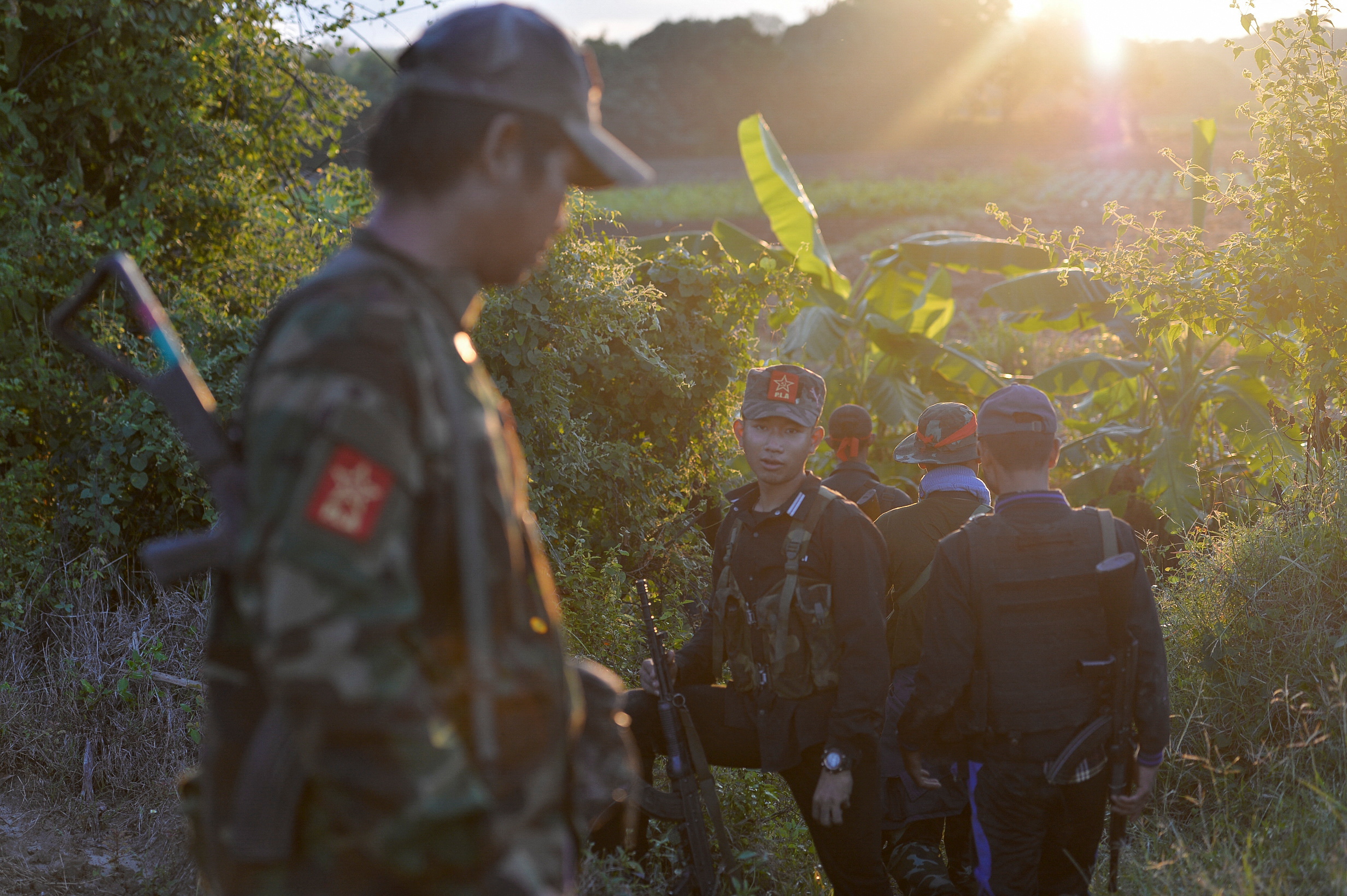 An anti-coup fighter in Sagaing region. He is walking with his comrades in a rural area. The sun is shining behind him.