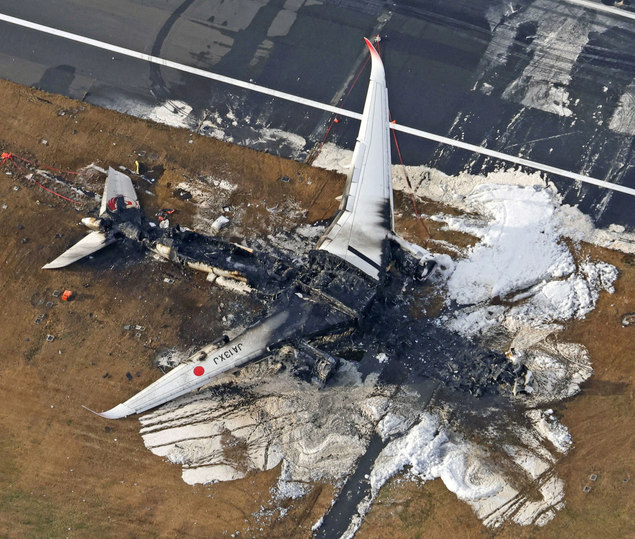 The burnt out wreckage of the JAL plane on the runway. The cabin in the middle is black and destroyed. The wings are still visible and white.