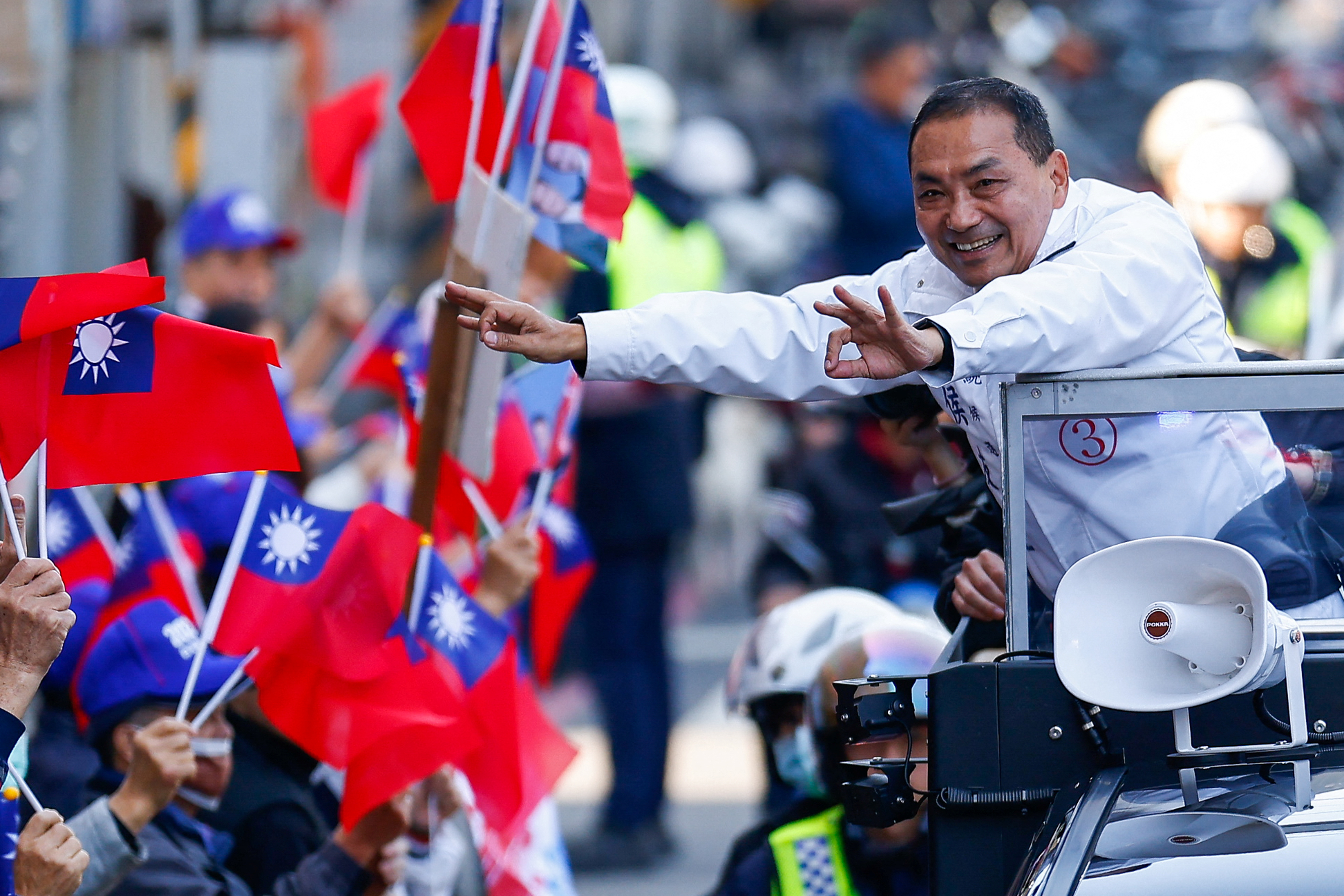 Hou Yu-ih, the KMT's candidate, reaching out to supporters at a campaign event. He's wearing a white jacket and smiling. They are waving Taiwan flags