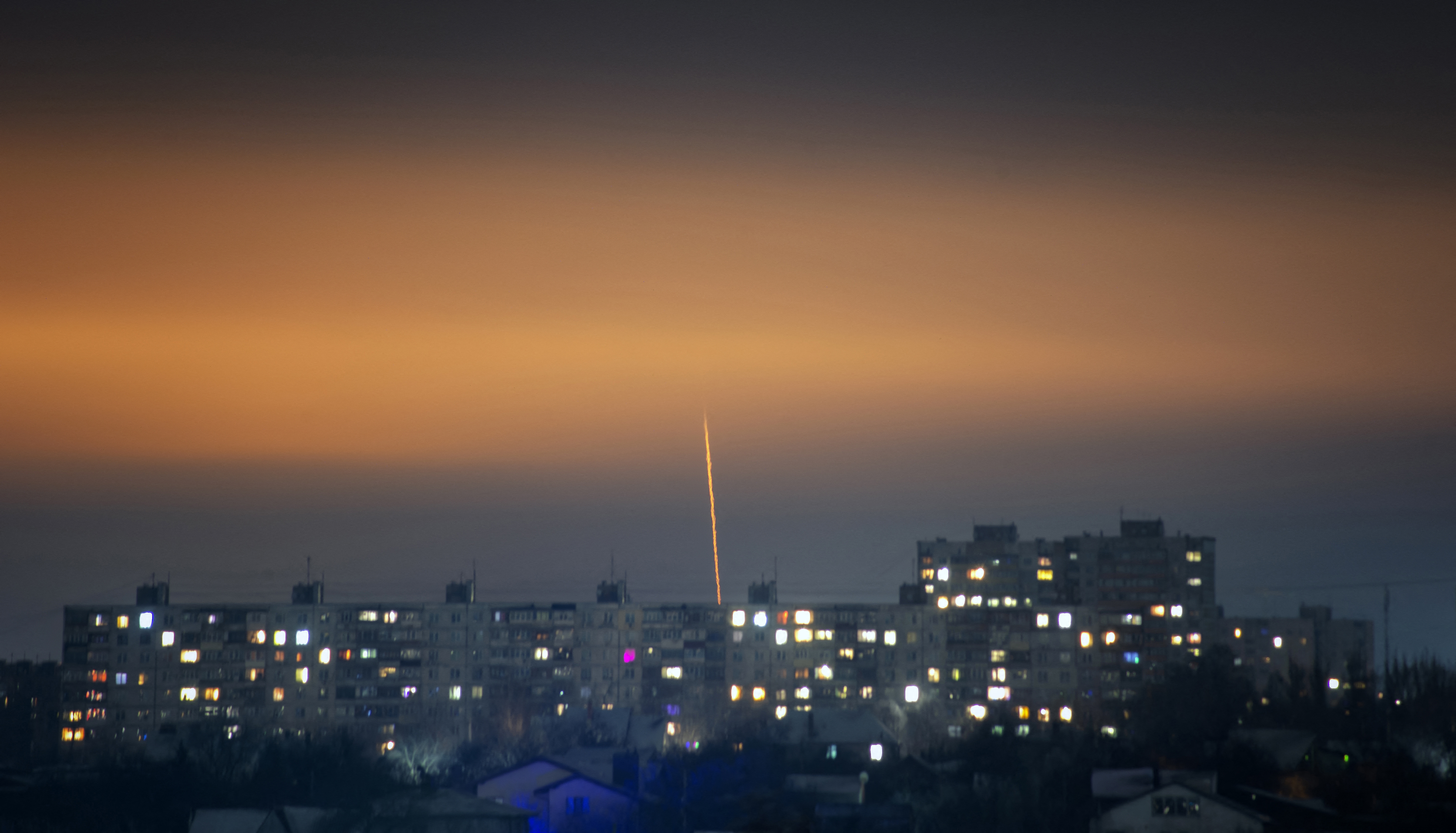 A missile launched from Belgorod in Russia towards the Ukrainian city of Kharkiv, The missile is leaving a trail of fire in the night sky. Buildings are illuminated below.