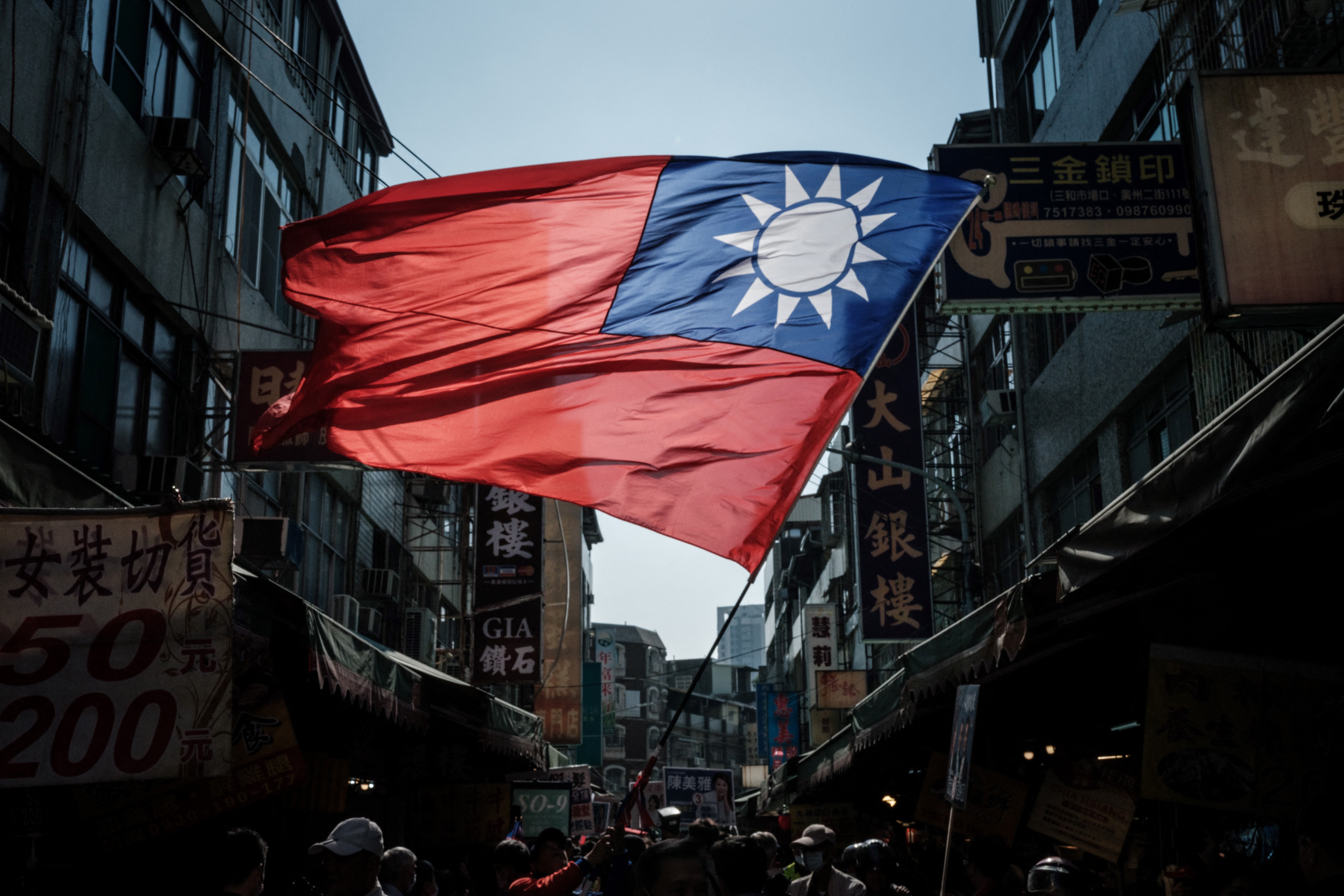 A Taiwan flag flown in the street. It's illuminated by the sky and there are buildings on eitehr side