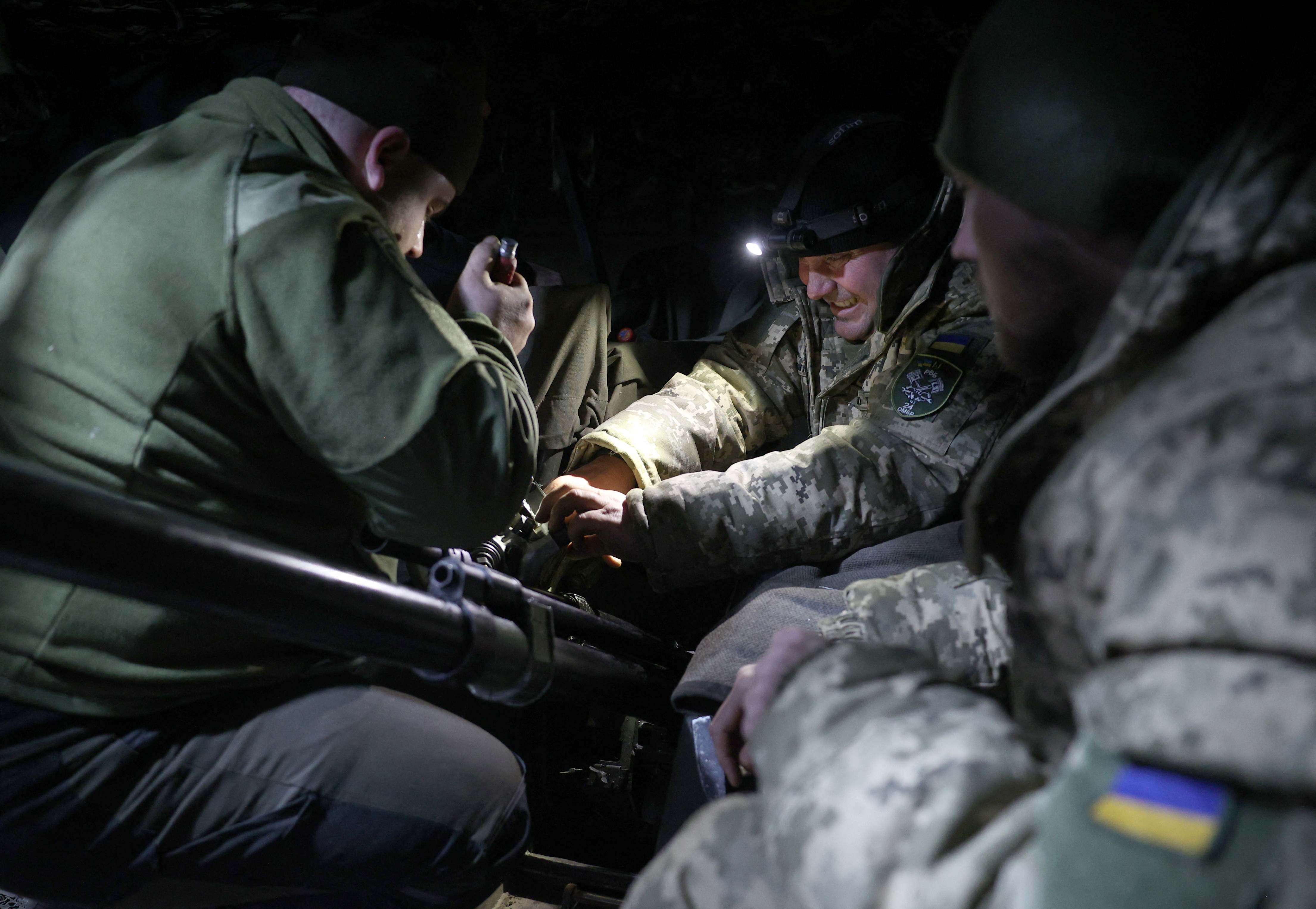 Ukrainian soldiers fitting an automatic cannon on an armoured military vehicle. They are huddled around the cannon. One is holding a torch.