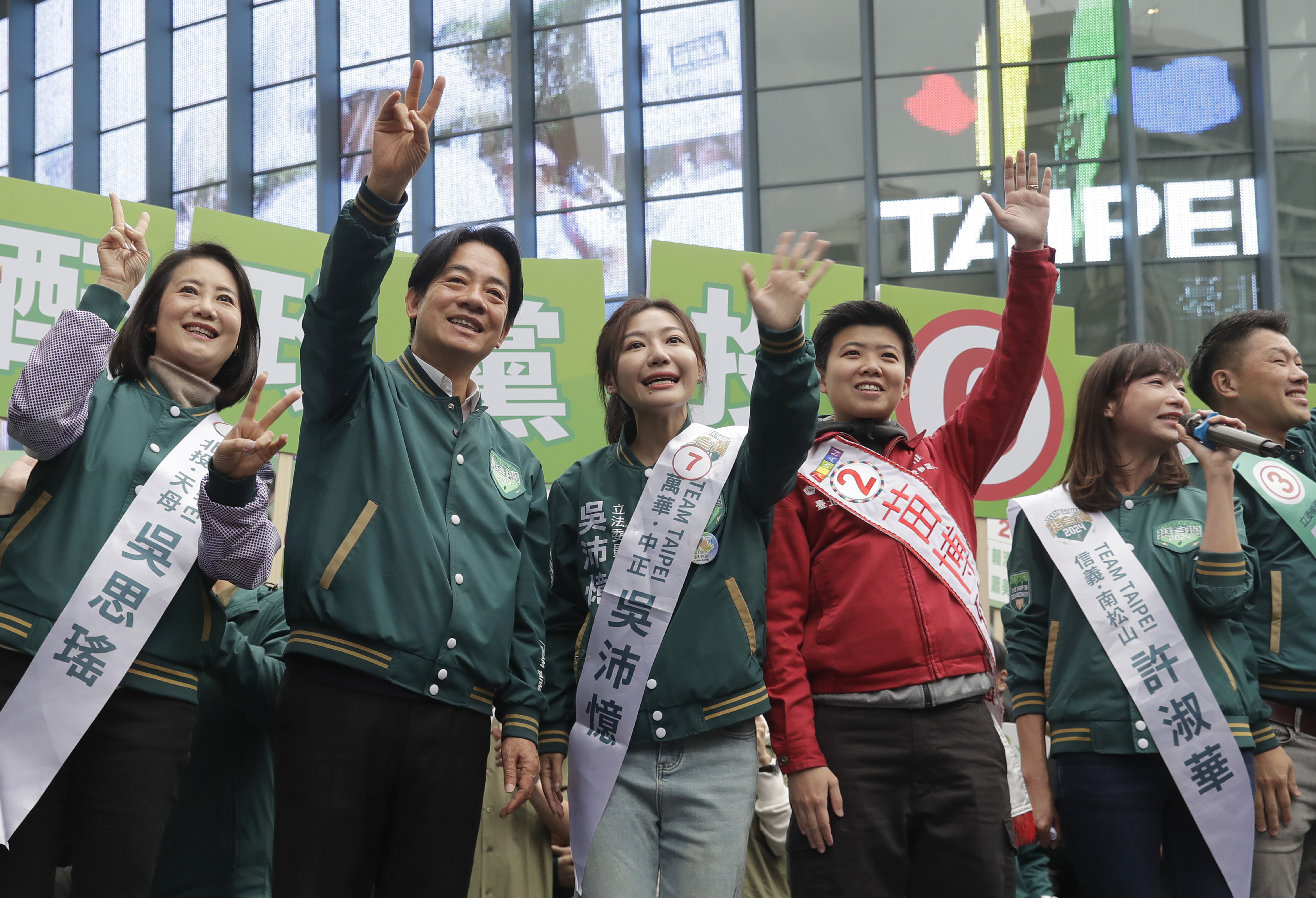 William Lai waving to the crowd. He is wearing a dark green baseball jacket and standing on a stage with some of the DPP's candidates for the legislature