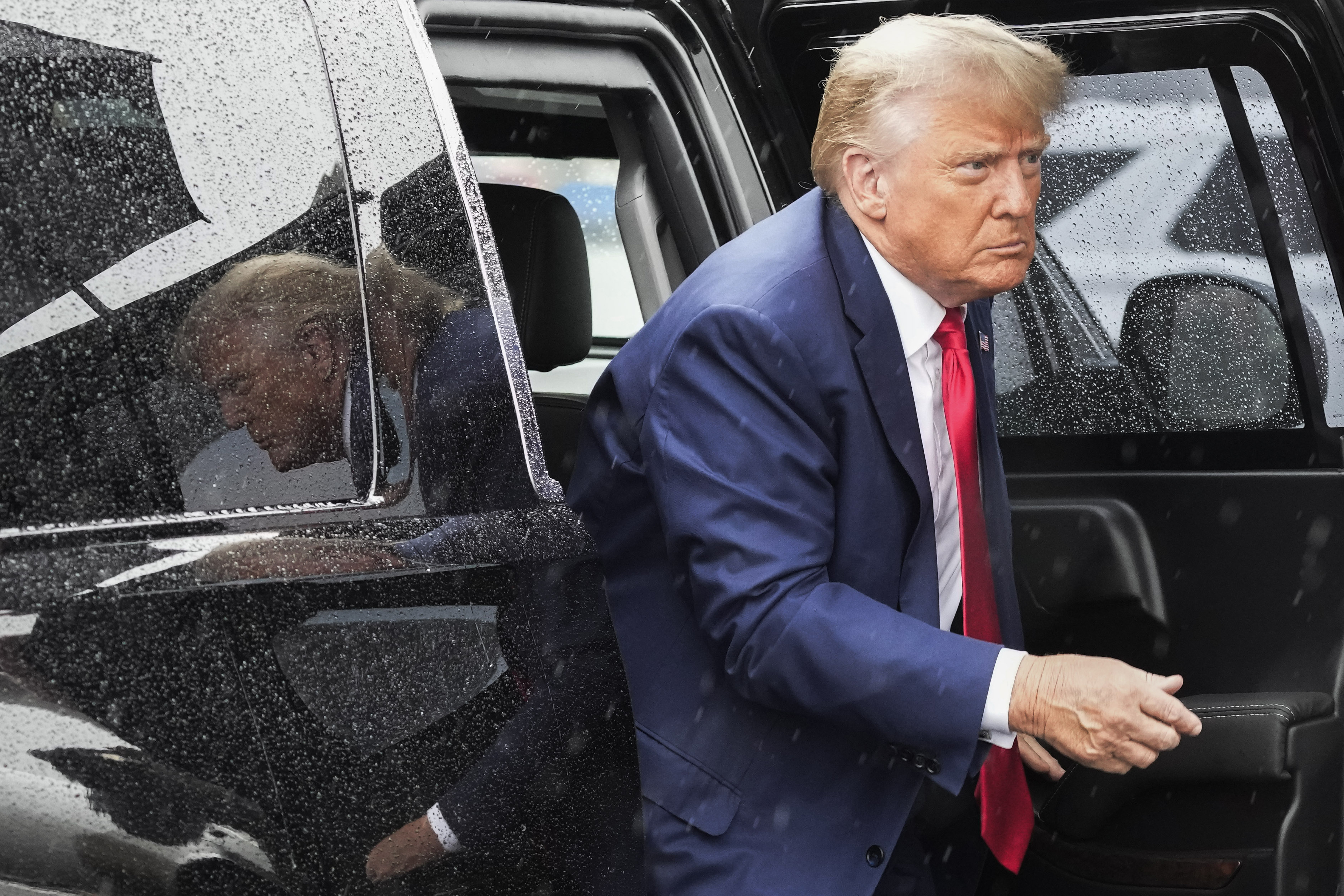 Donald Trump steps out of a black SUV vehicle, wearing a blue suit and red tie.