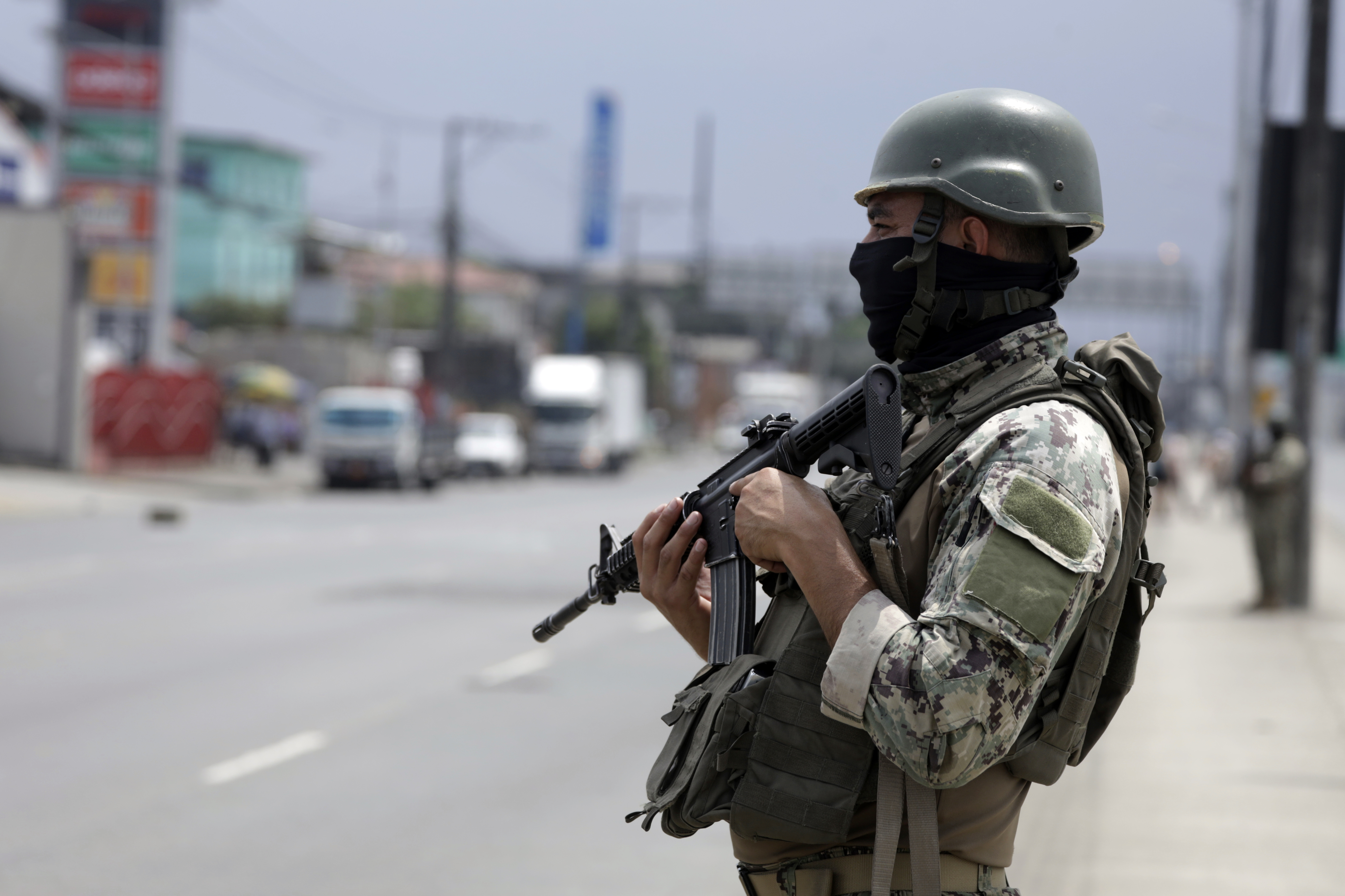 A soldier in a helmet, flak jacket and fatigues stands on a city street, holding a gun.