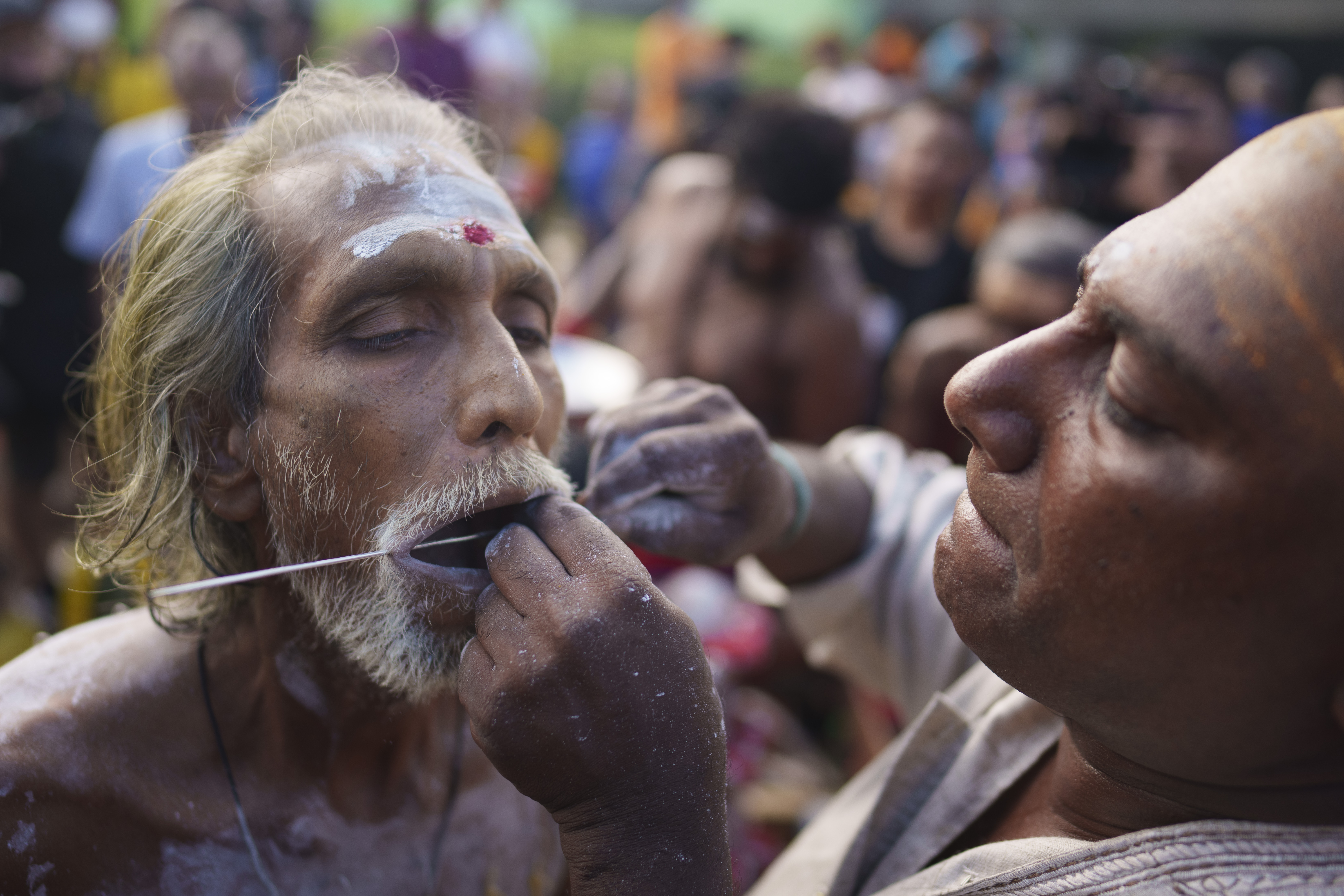 A man with a white beard and moustache having his tongue and cheeks pierced with a metal rod.
