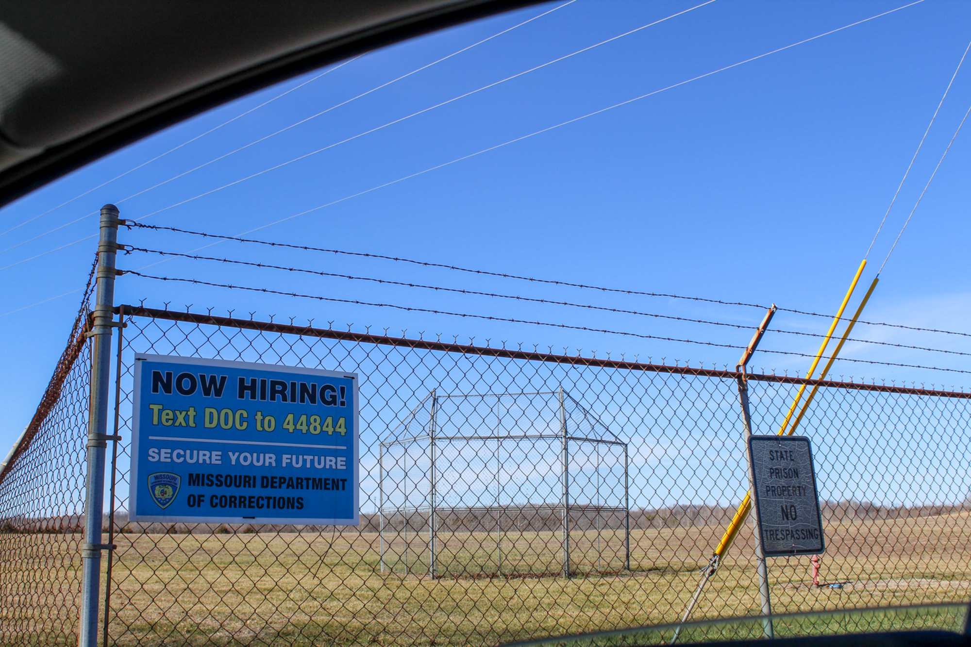 A recruitment sign on a wire fence outside a prison says the prison is recruiting corrections officers.