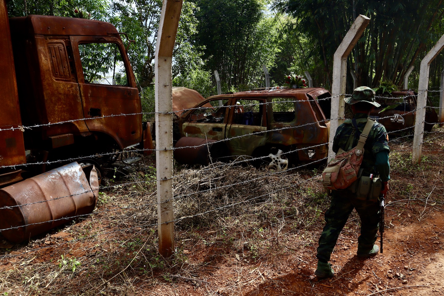 A KNPLF soldier walks past the massacre site. The vehicles are rusty and behind a barbed wire fence. There are trees around.