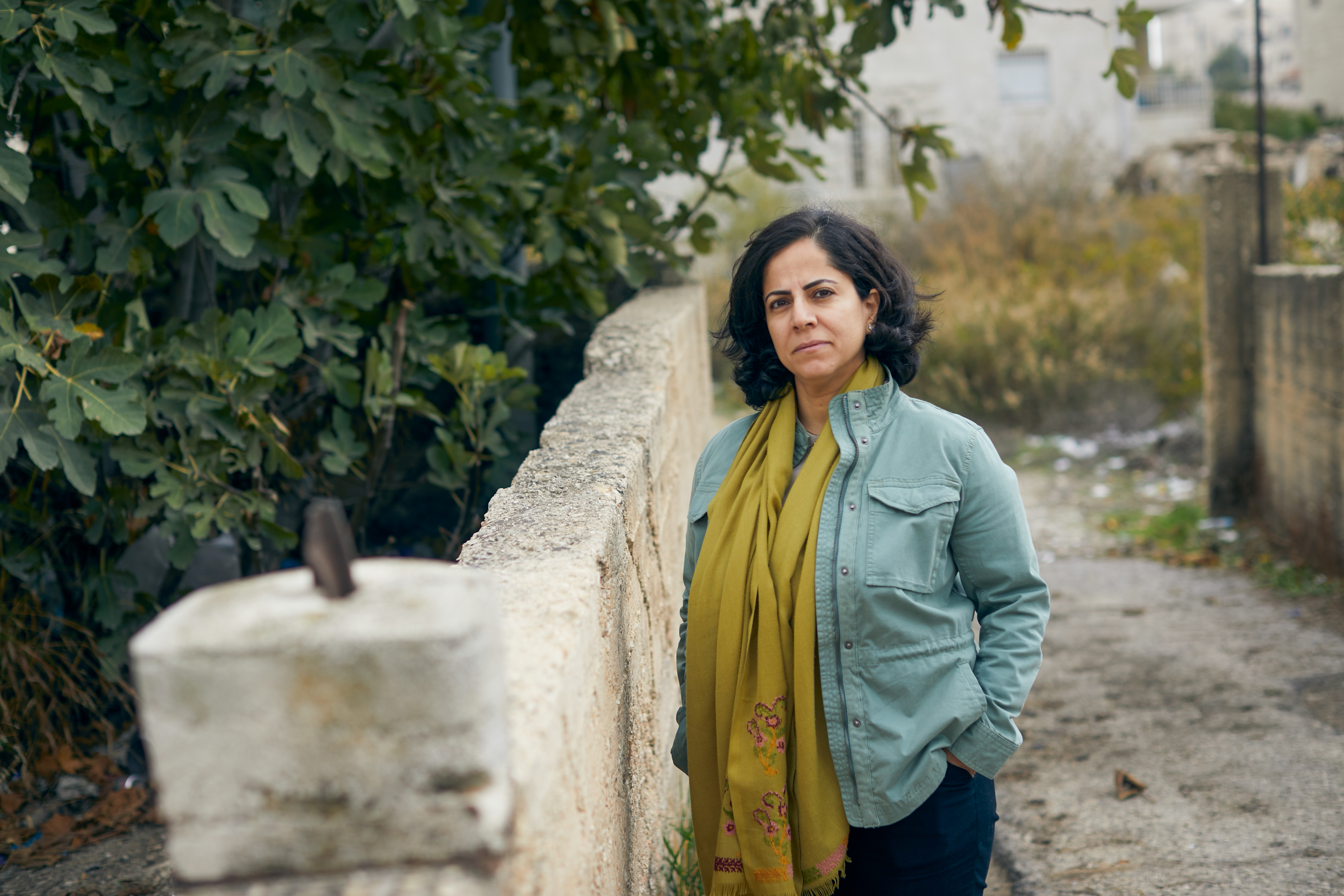 Erab in a blue shirt stands next to a stone wall in a garden with her hands in her pockets