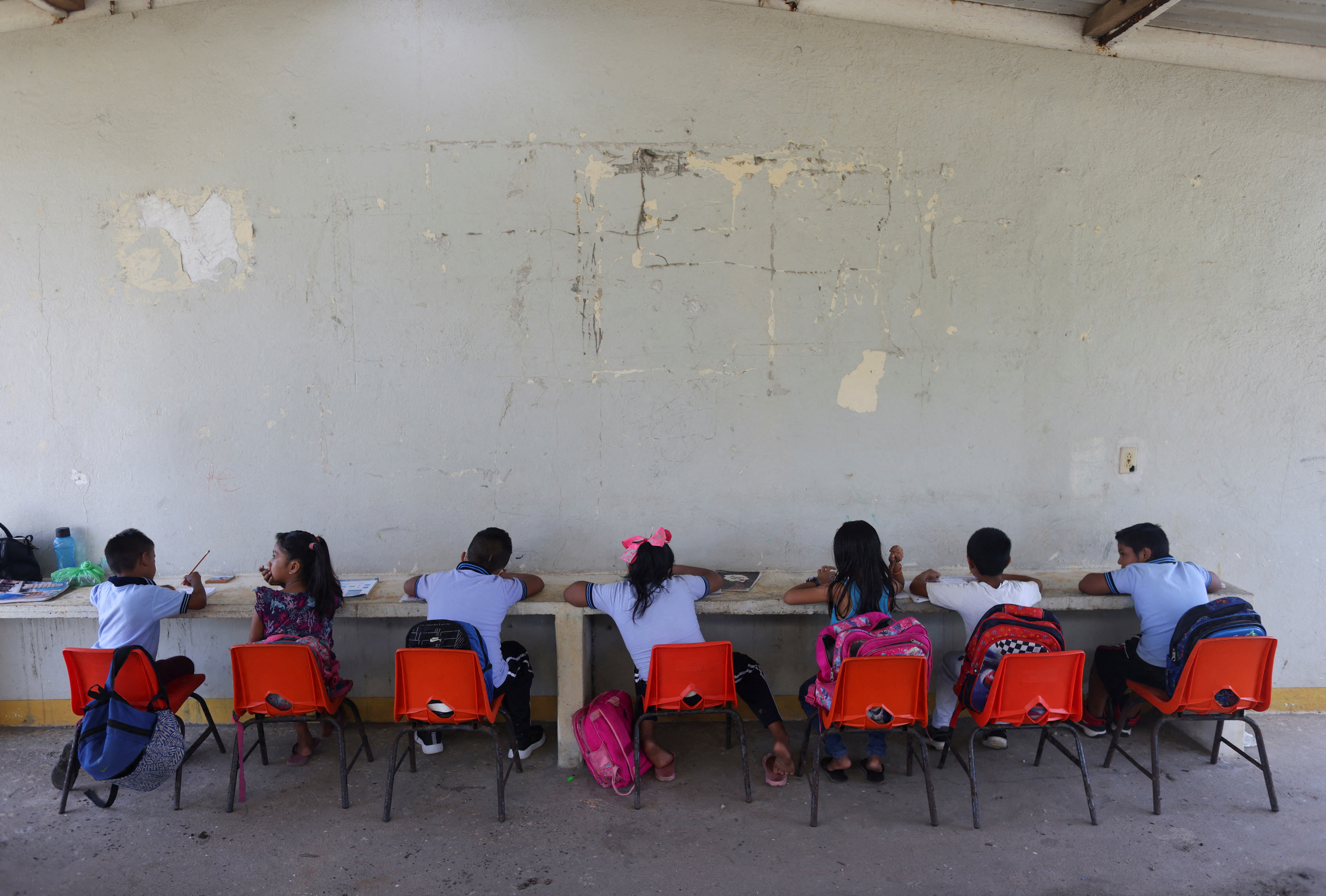 A row of young pupils sit in orange plastic chairs at a long desk placed against a peeling wall in a Mexico school.