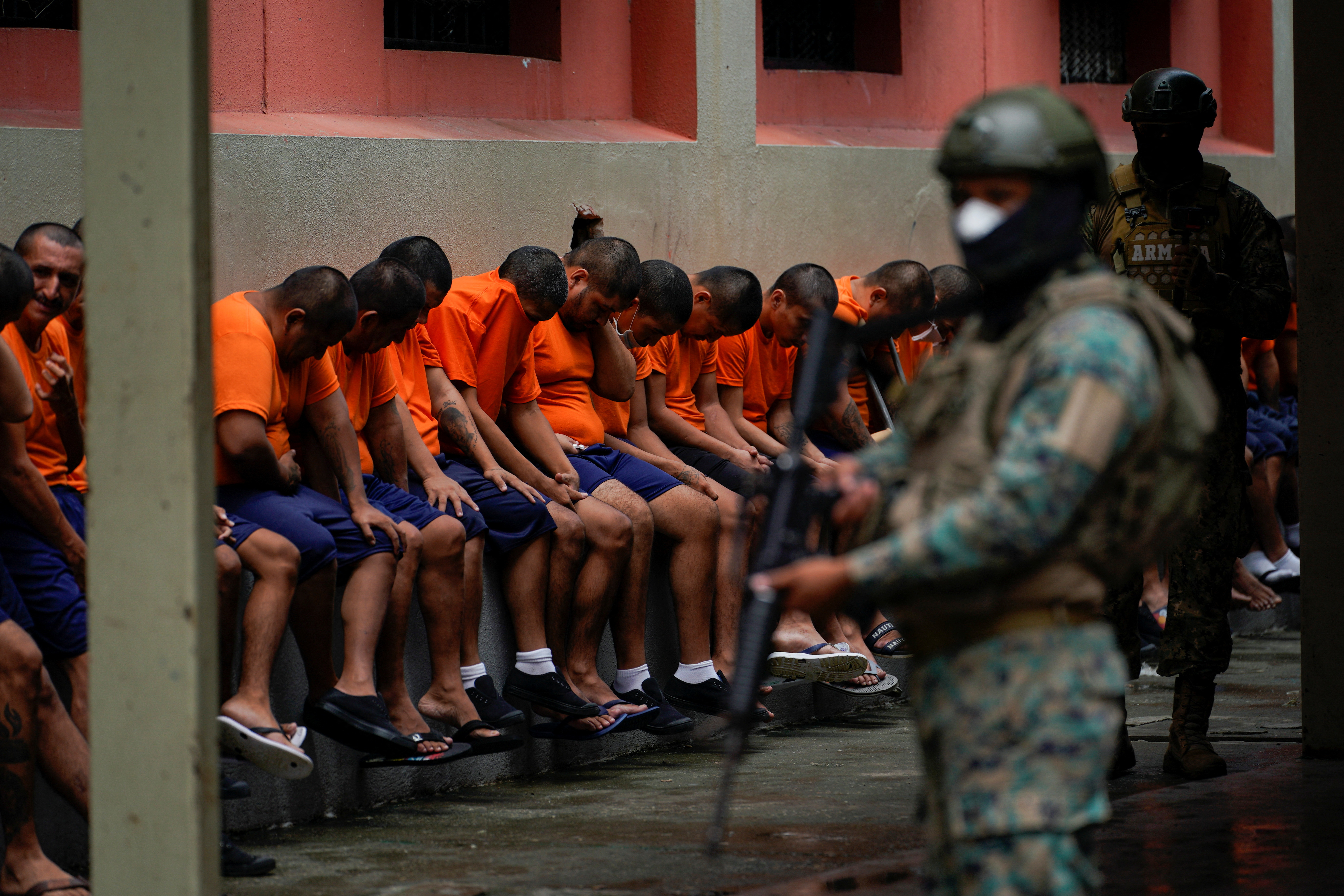 A row of prisoners, clad in orange shirts, sit with their heads bowed on a long, crowded bench. In the foreground, a military member in full gear — helmet, face mask, camouflage and a gun — stands guarding them.