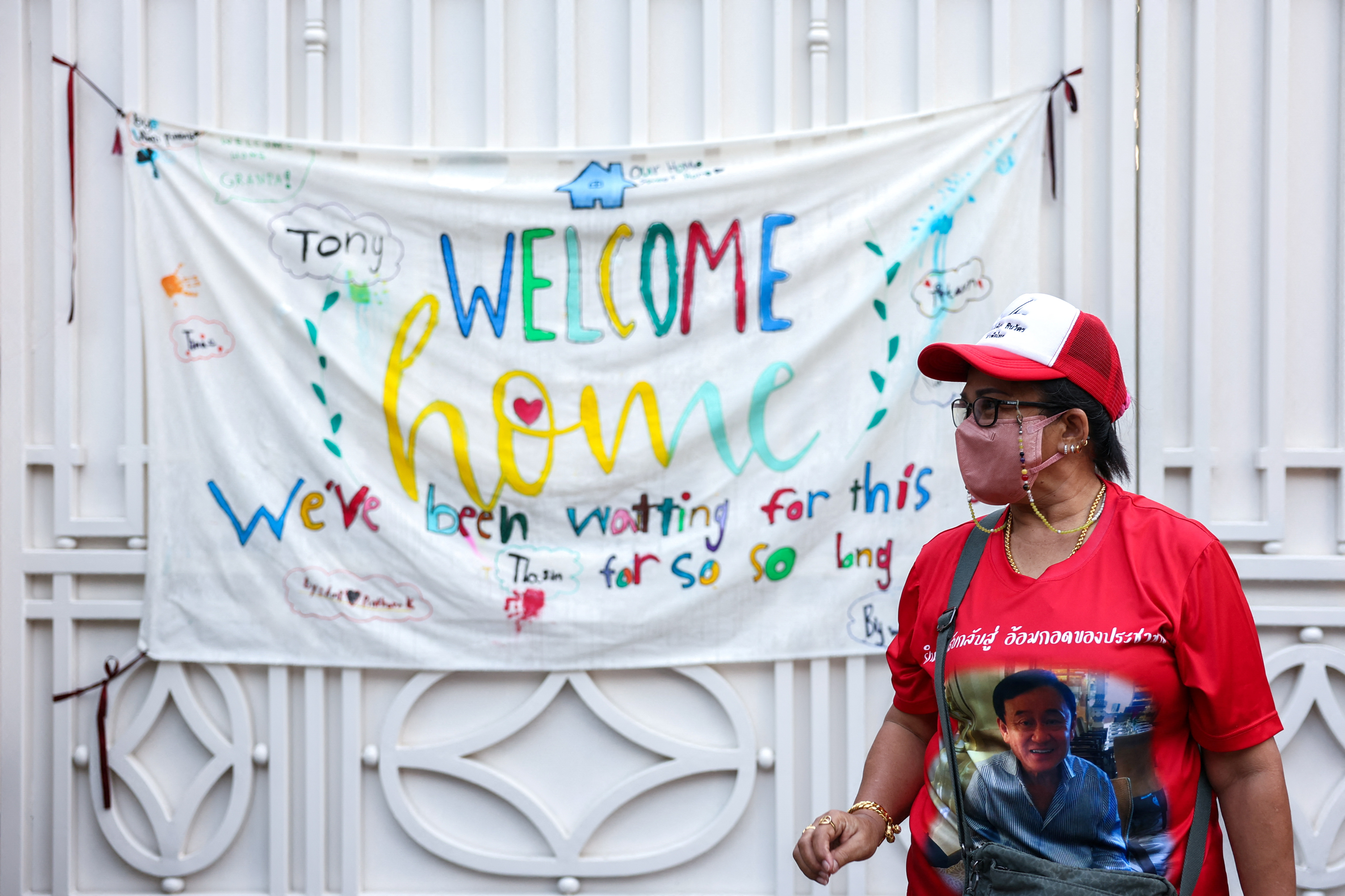 A supporter in a red t-shirt with Thaksin's picture on the front, and wearing a red and white cap, standings in front of a banner that says, 'Welcome home. We've been waiting for this for so so long'.