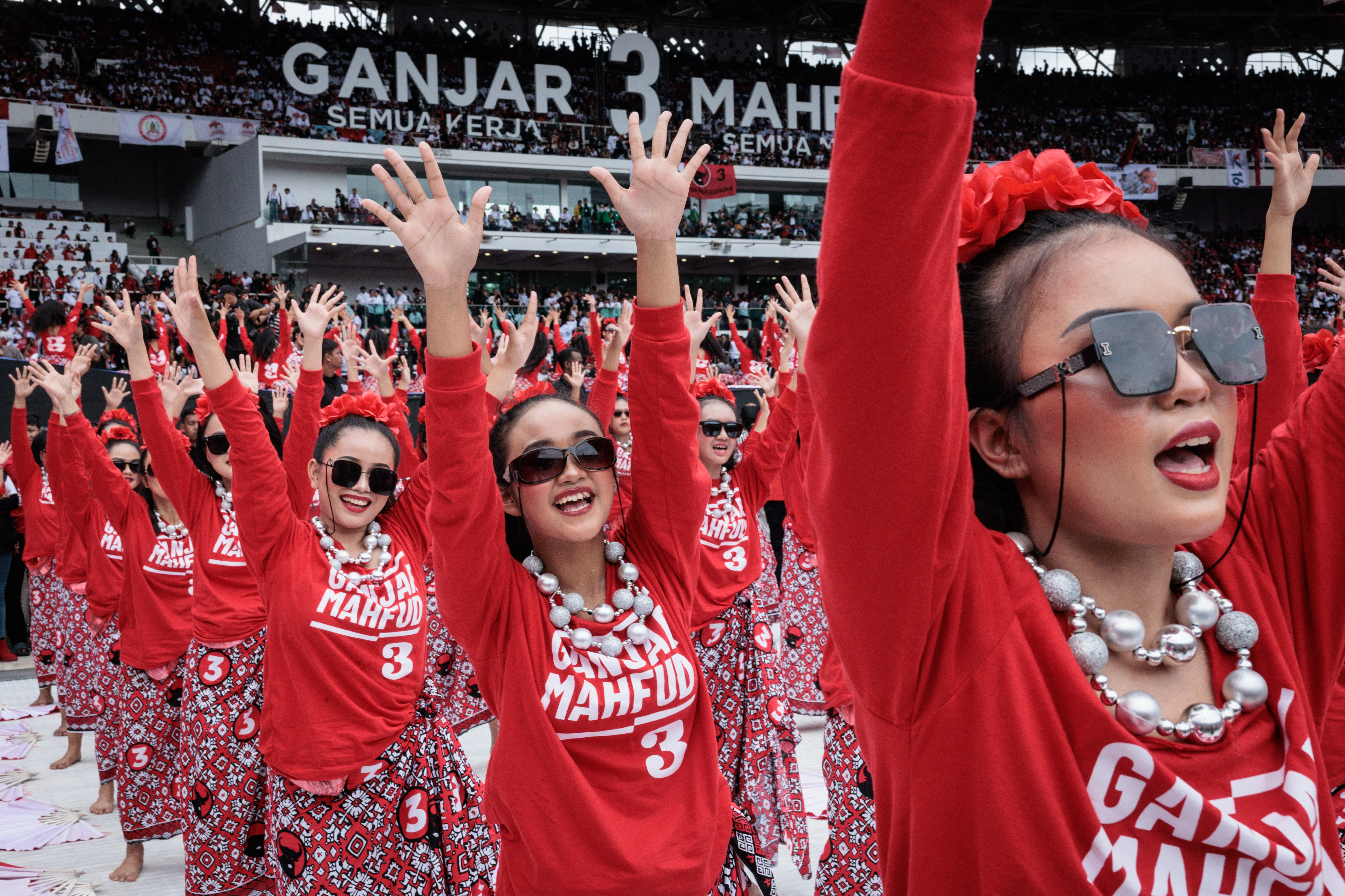 Women dressed in red and white dance at a campaign rally for Ganjar Pranowo. They have their hands in the air.