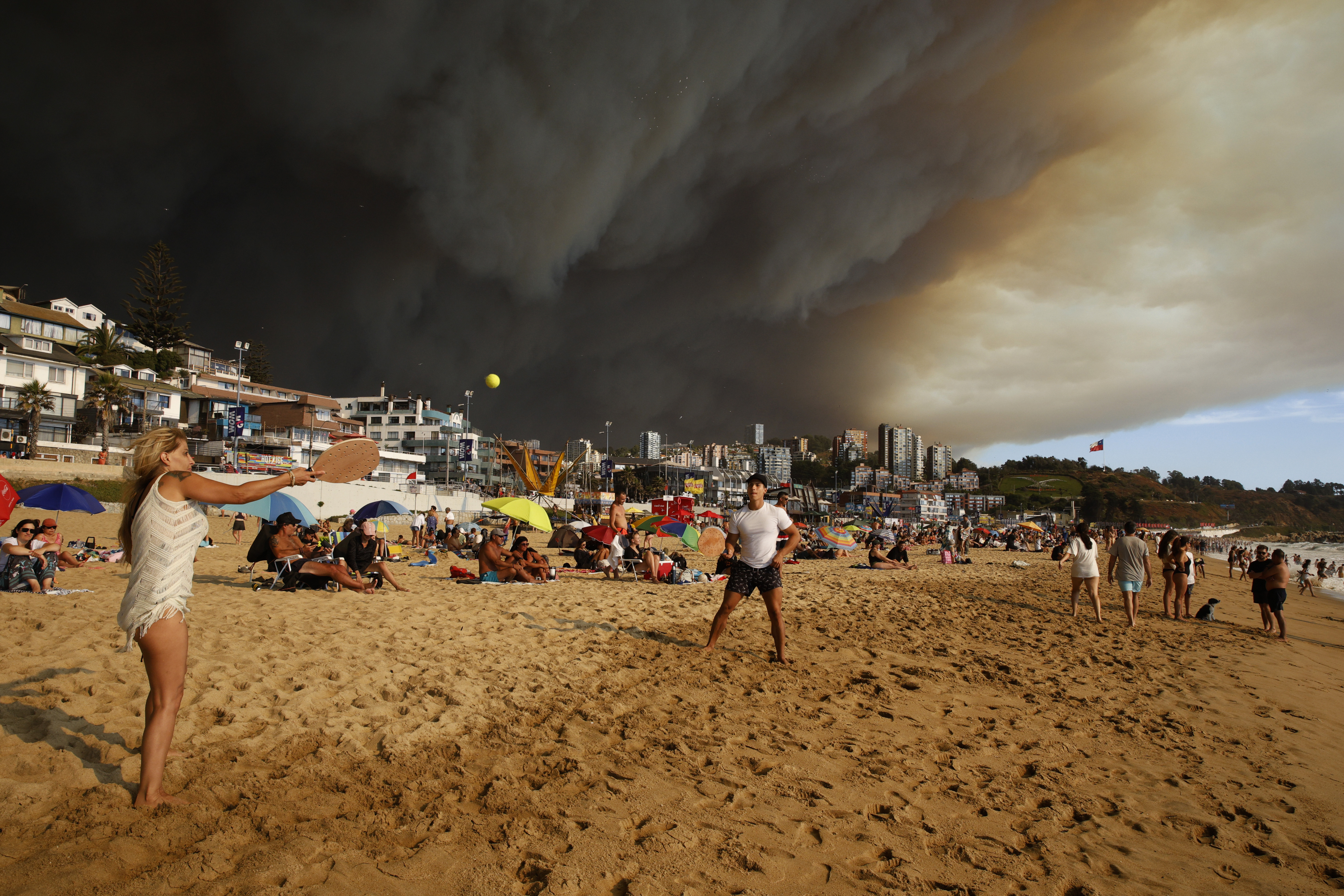 Vacationers play paddle ball on a beach backdropped by a darkening sky caused by smoke from nearby forest fires, in Viña del Mar, Chile, Friday, Feb. 2