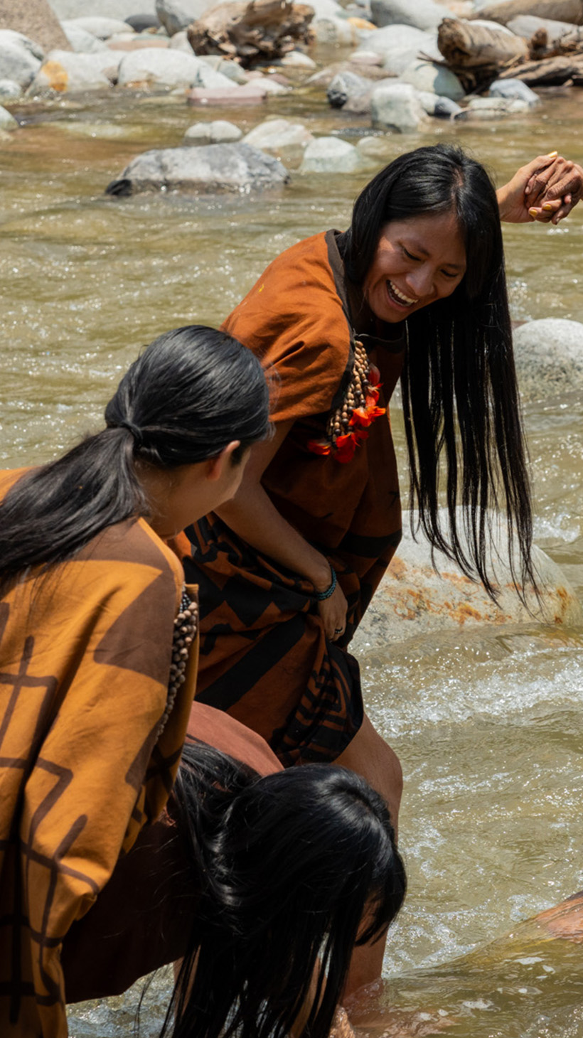 Three Indigenous Ashaninka women stand and smile in a river lined with rocks.