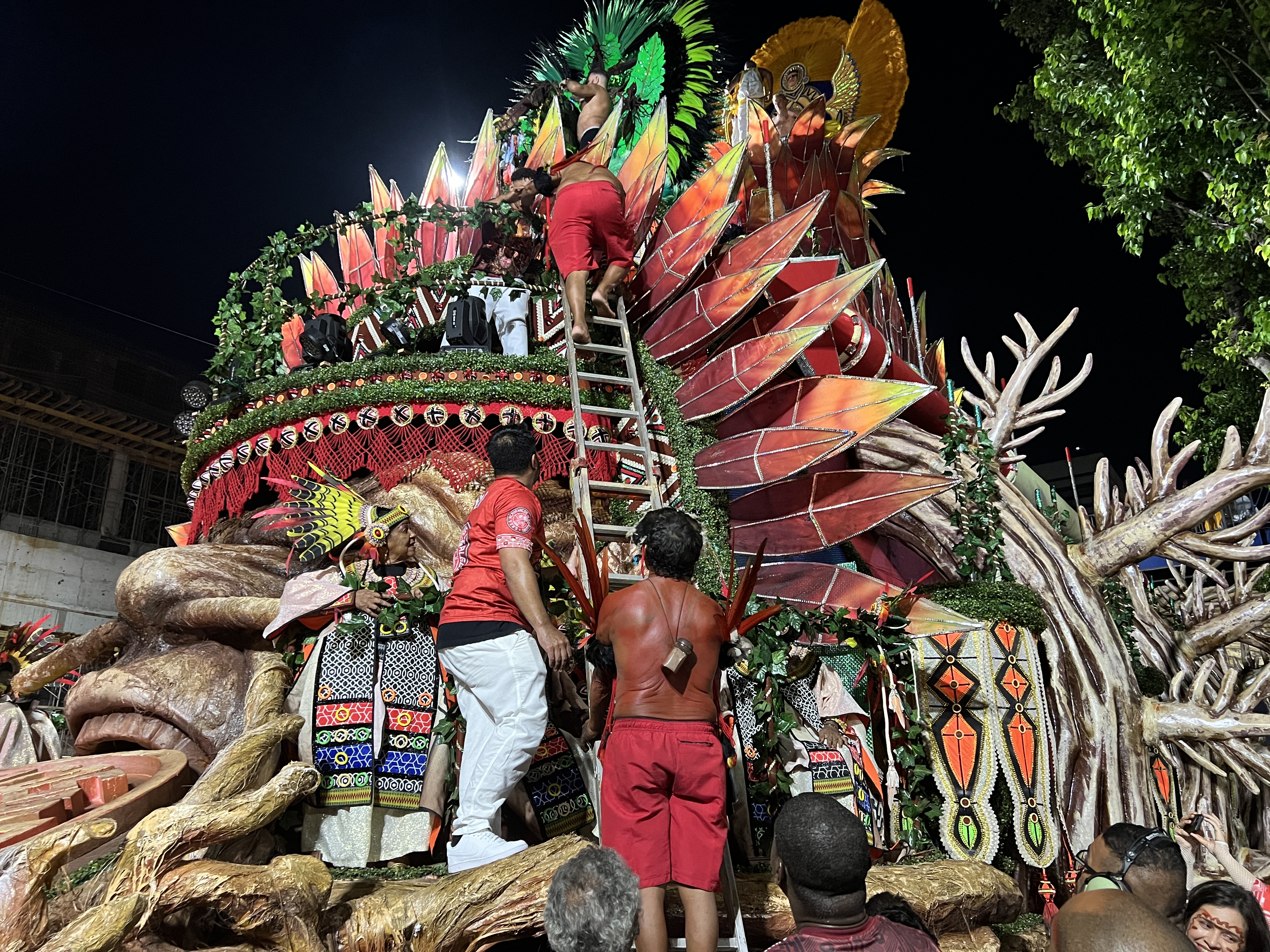 A parade participant at Rio de Janeiro's Sambadrome holds a metal ladder while another person climbs to the top of a parade float, designed to appear as if an Indigenous face is emerging from wood.