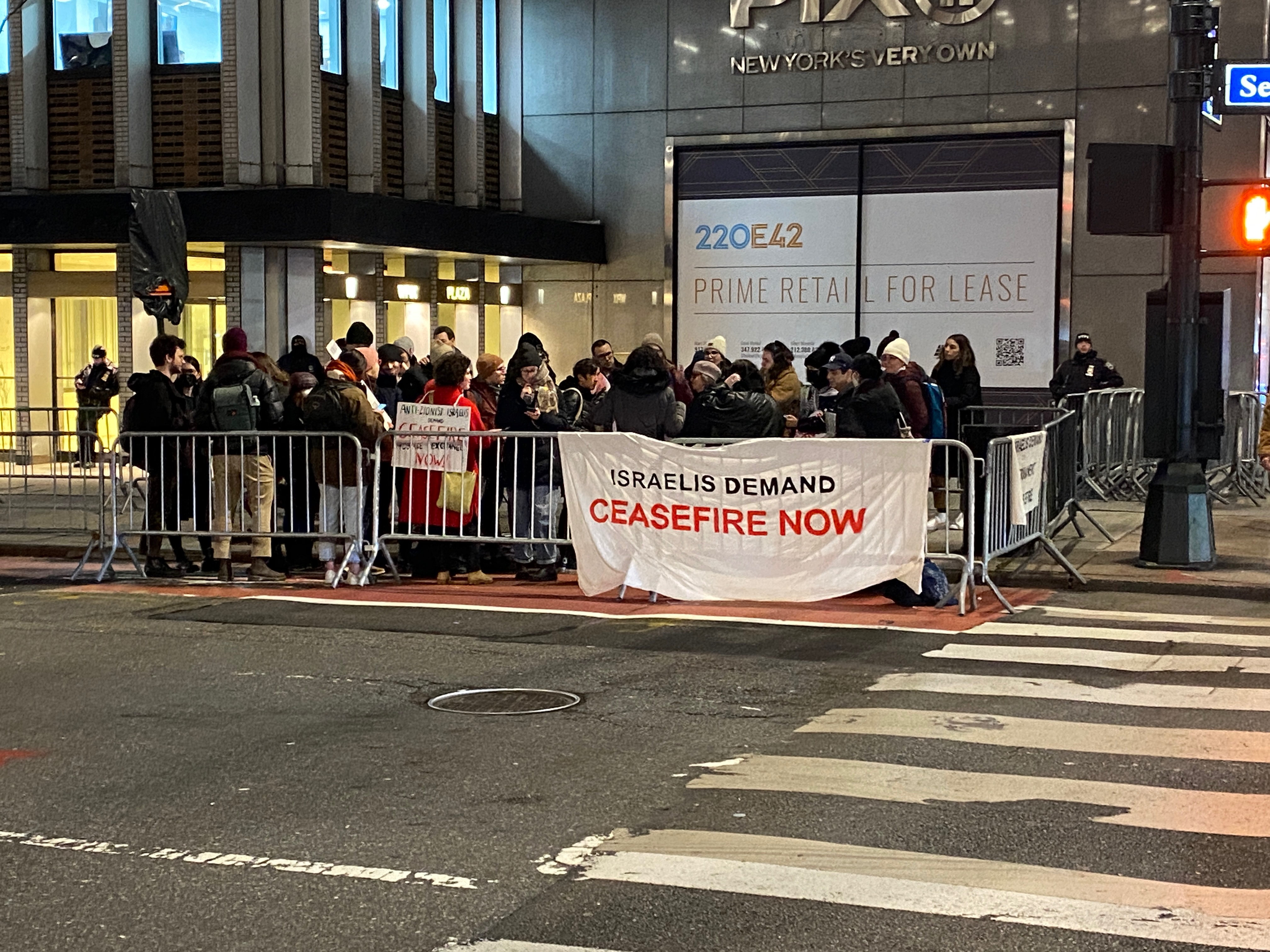 Protesters gather on a street corner with a banner that reads, "Israelis demand ceasefire now."