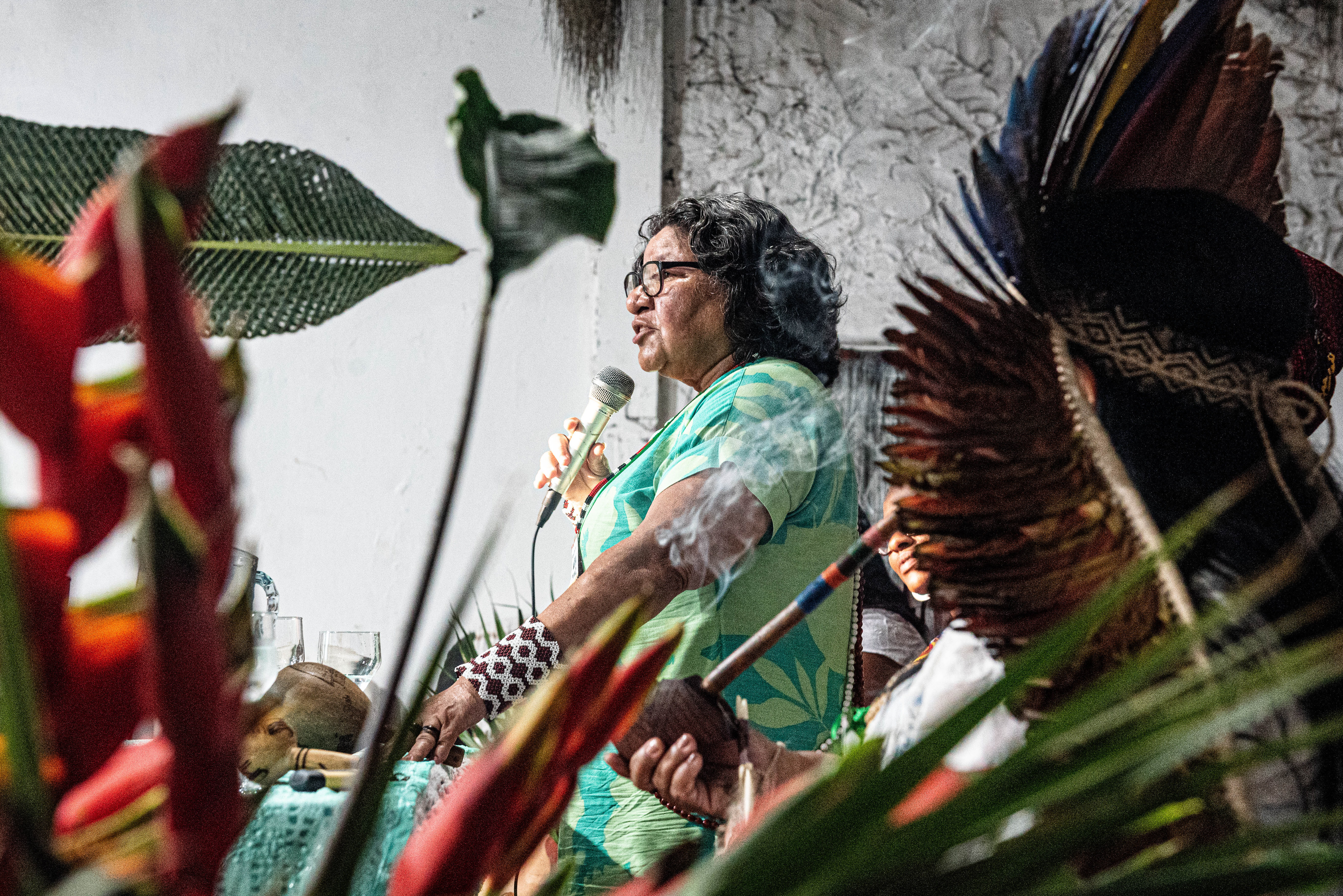 A photo of Maria de Fátima Muniz speaking into a microphone, seen through a space between palm fronds.