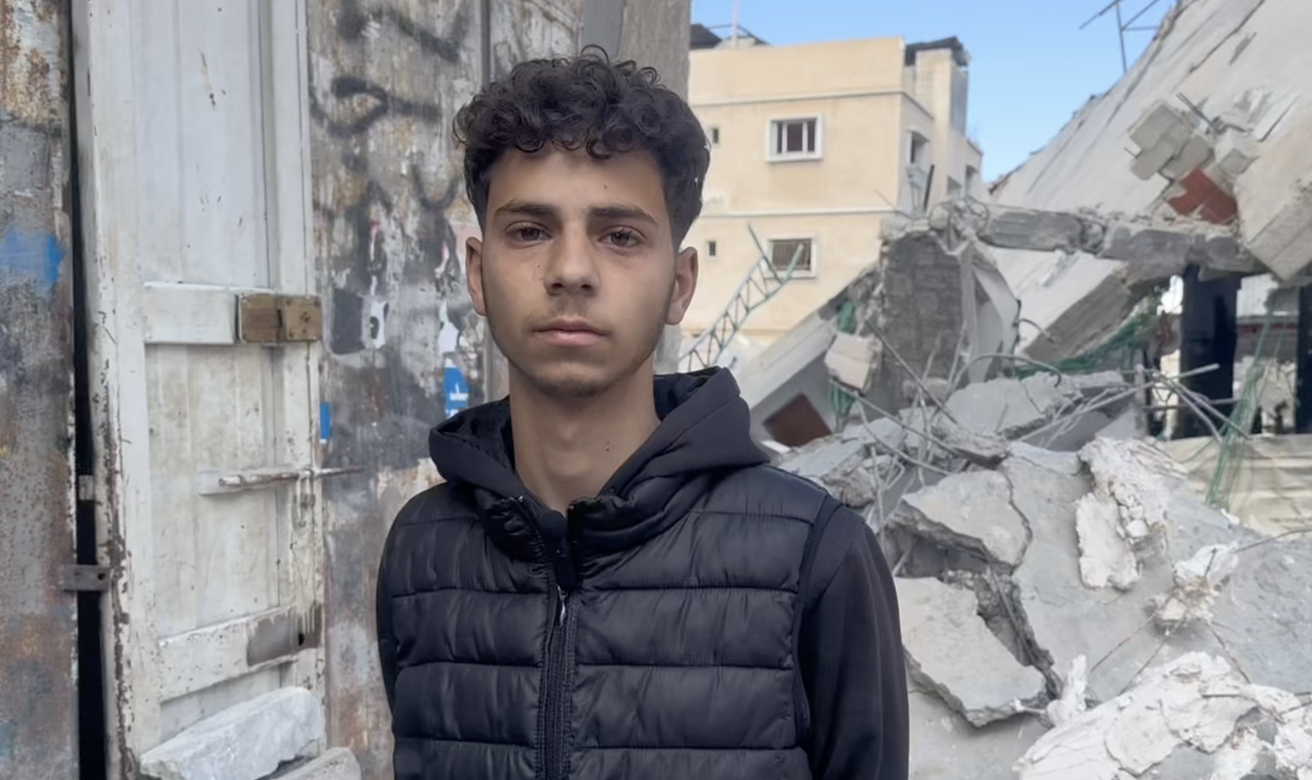 Shehab Omar Abu al-Hanud stands with destruction behind him, holding back his tears