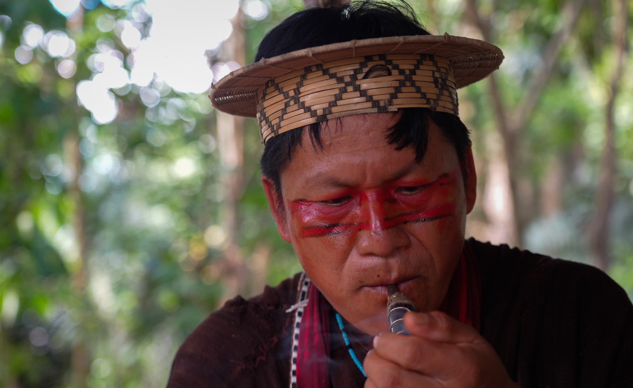 Tsitsiri Samaniego, wearing a straw headdress, lights a pipe and smokes it.