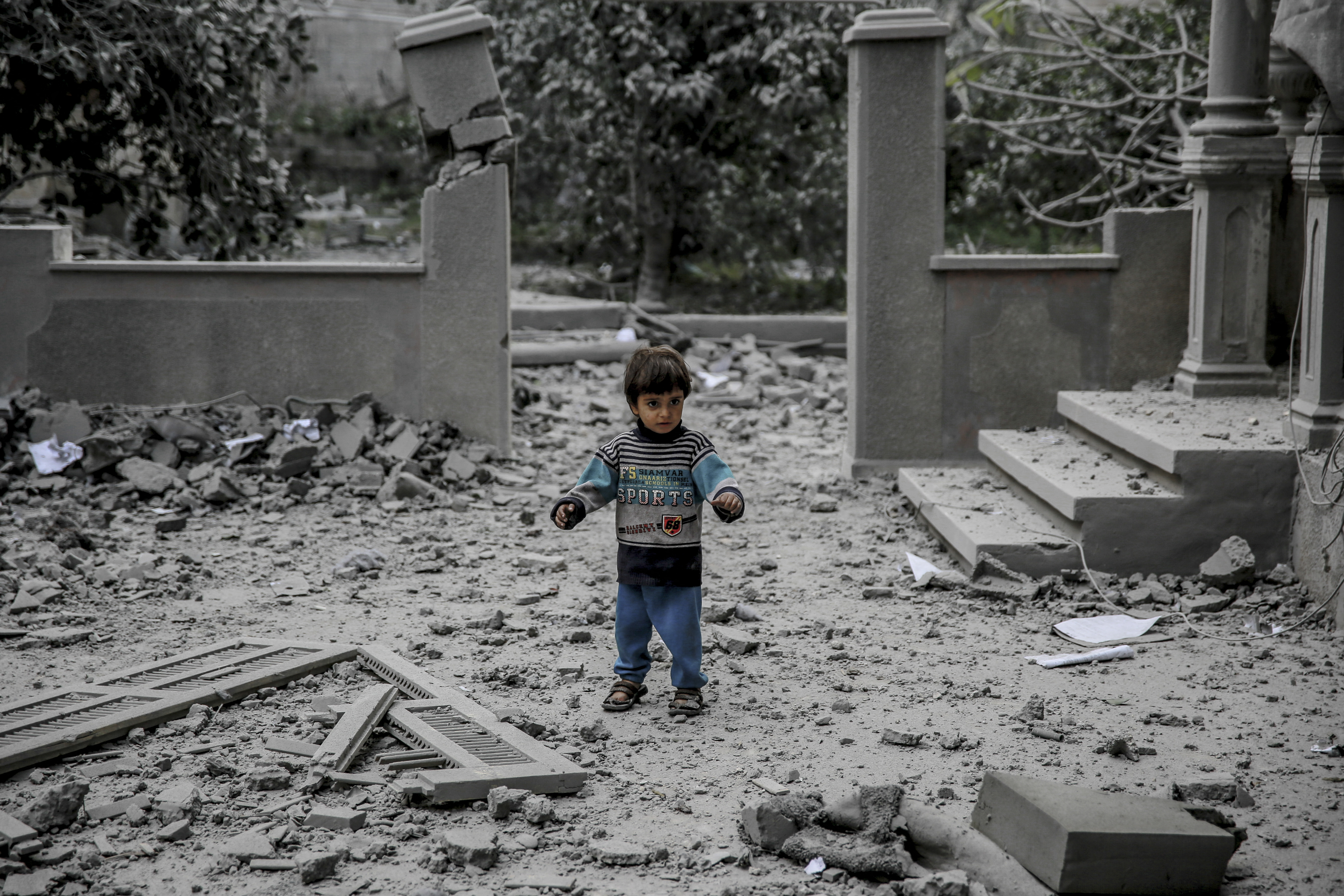 a small toddler stands in front of a destroyed building covered in grey dust