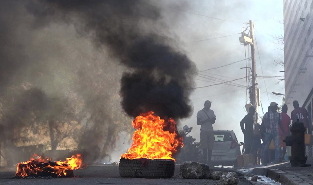 Flames and smoke rise from a pile of tyres set alight near Port au Prince's main prison. People can be seen through the smoke.