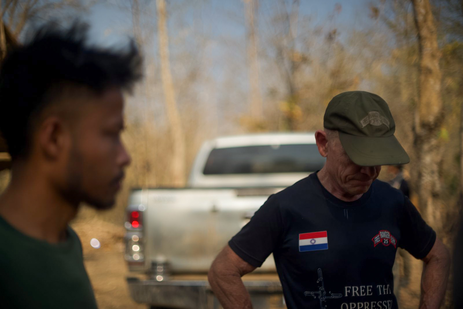 Dave Eubank of the Free Burma Rangers. He's wearing an FBR shirt and has his hands on his hips looking down at the ground. A pick-up truck is behind him, Another man is standing to the left