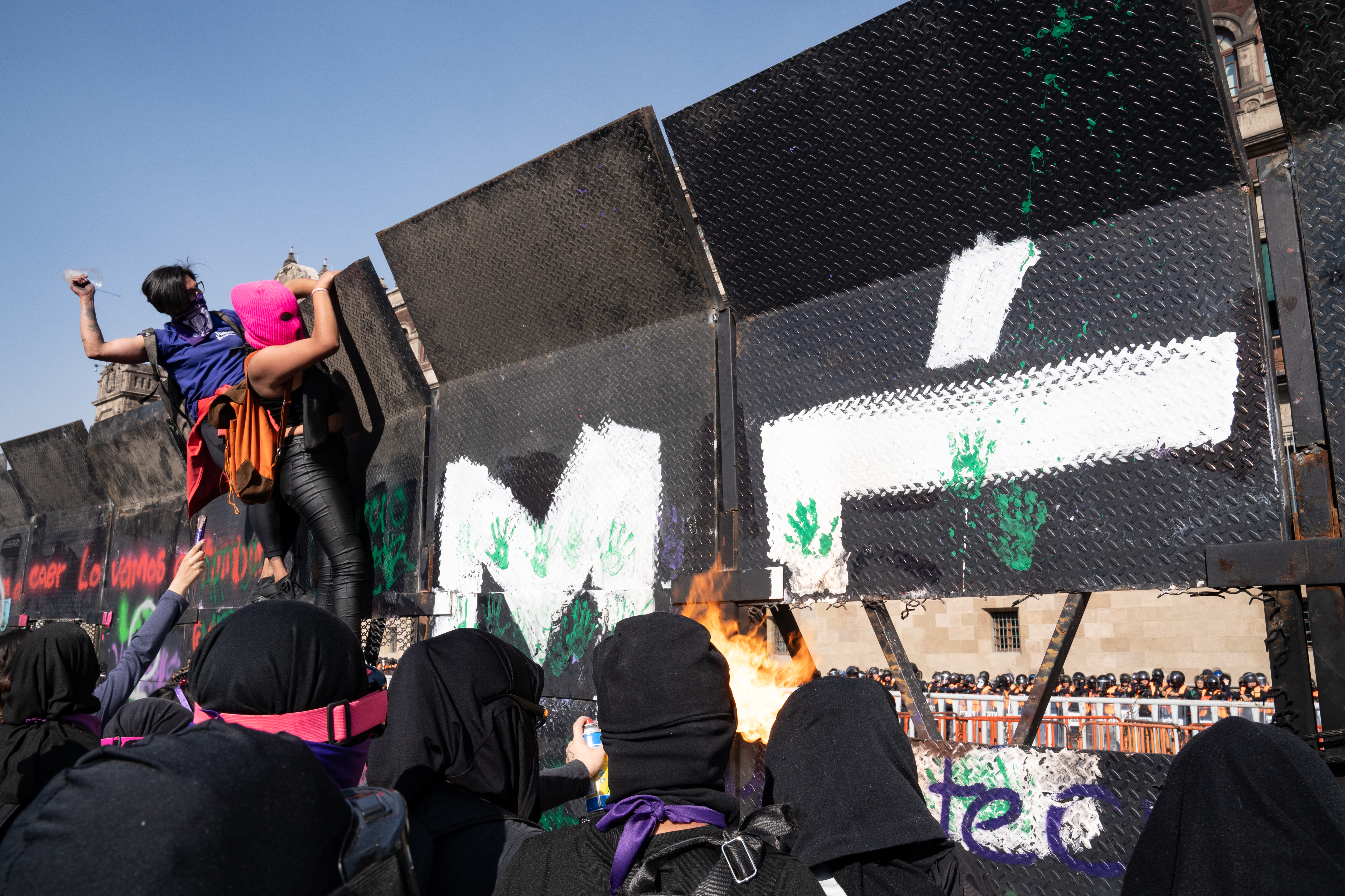 Hundreds of police stand far behind metal barriers as protesters shout at them and throw light objects such as plastic cups or other rubbish. Some protesters used spray paints to start fires near the metal barriers, burning banners or pieces of cardboard [Lexie Harrison-Cripps / Al Jazeera]
