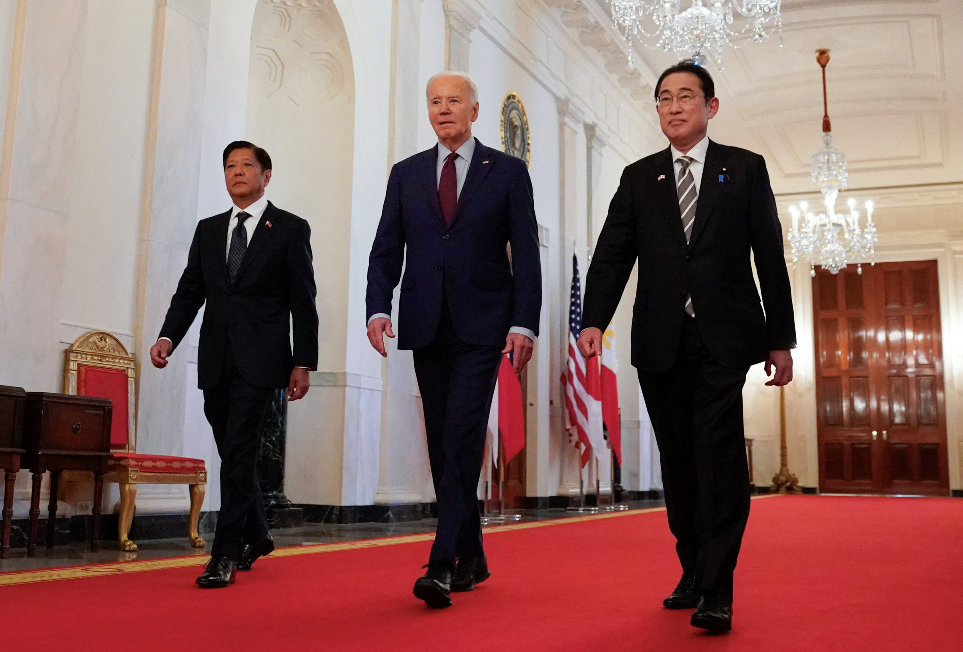 US President Joe Biden escorts Philippines President Ferdinand Marcos Jr and Japanese Prime Minister Fumio Kishida to their trilateral summit at the White House. They're walking along a corridor on a red carpet. They're all wearing dark suits and look relaxed.