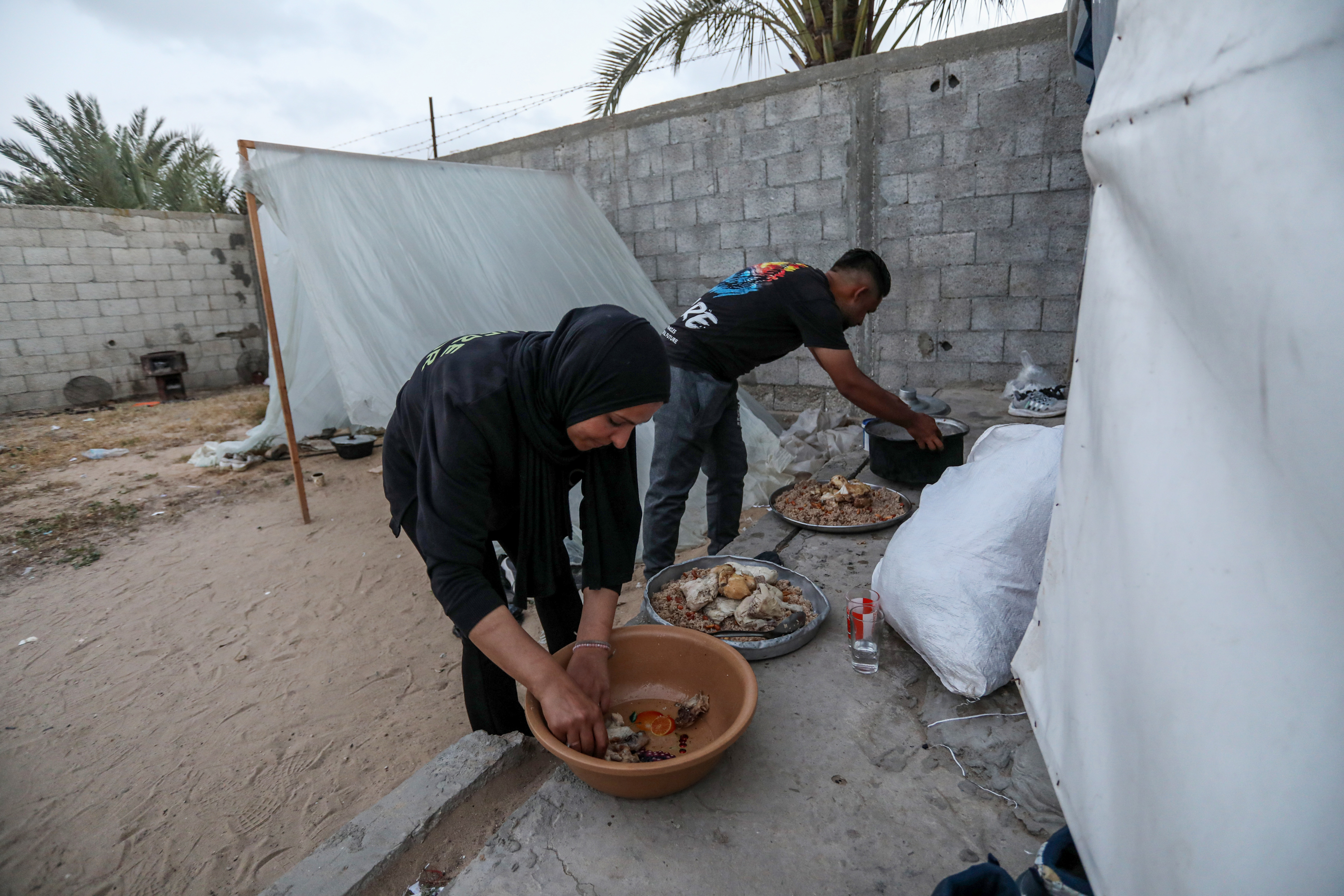 Siham is helped by a relative as she sets pots of food on a sandy bench to serve out into platters