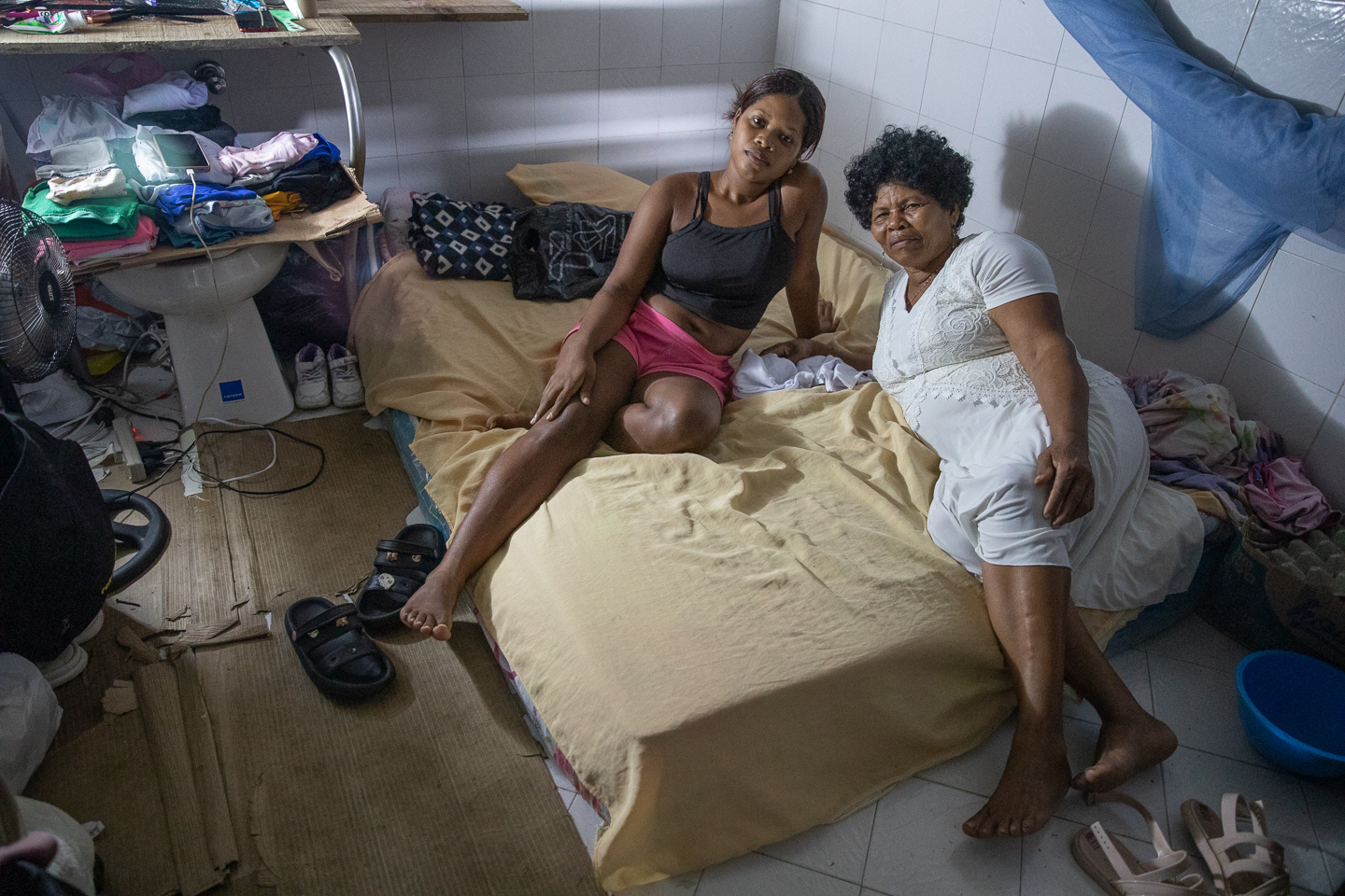 Two women lie on a mattress plopped on the floor of a coliseum.