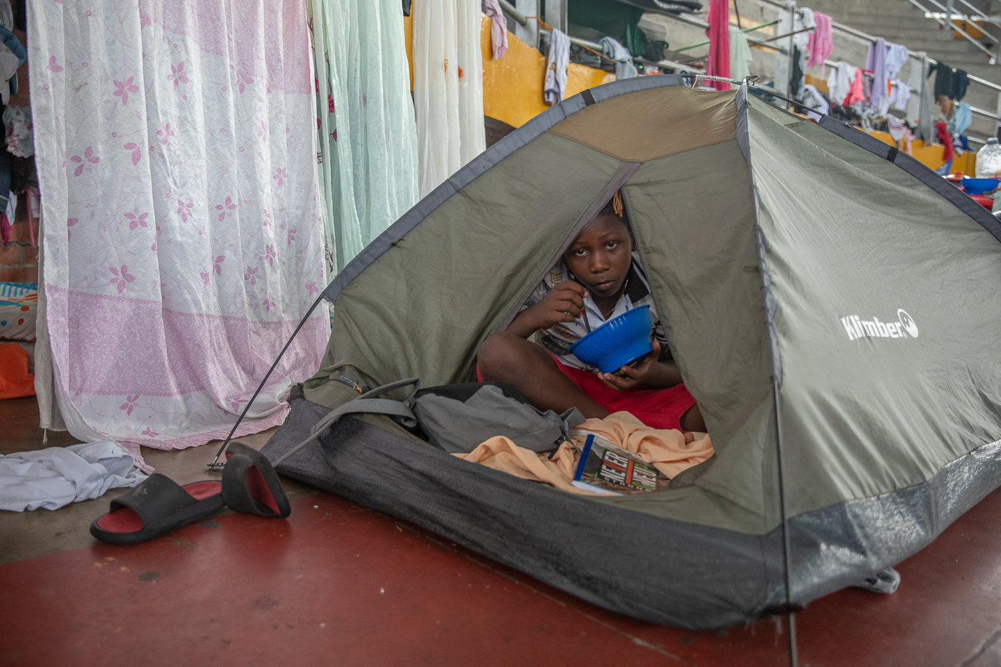 A child eating from a bowl peers out from a tent.