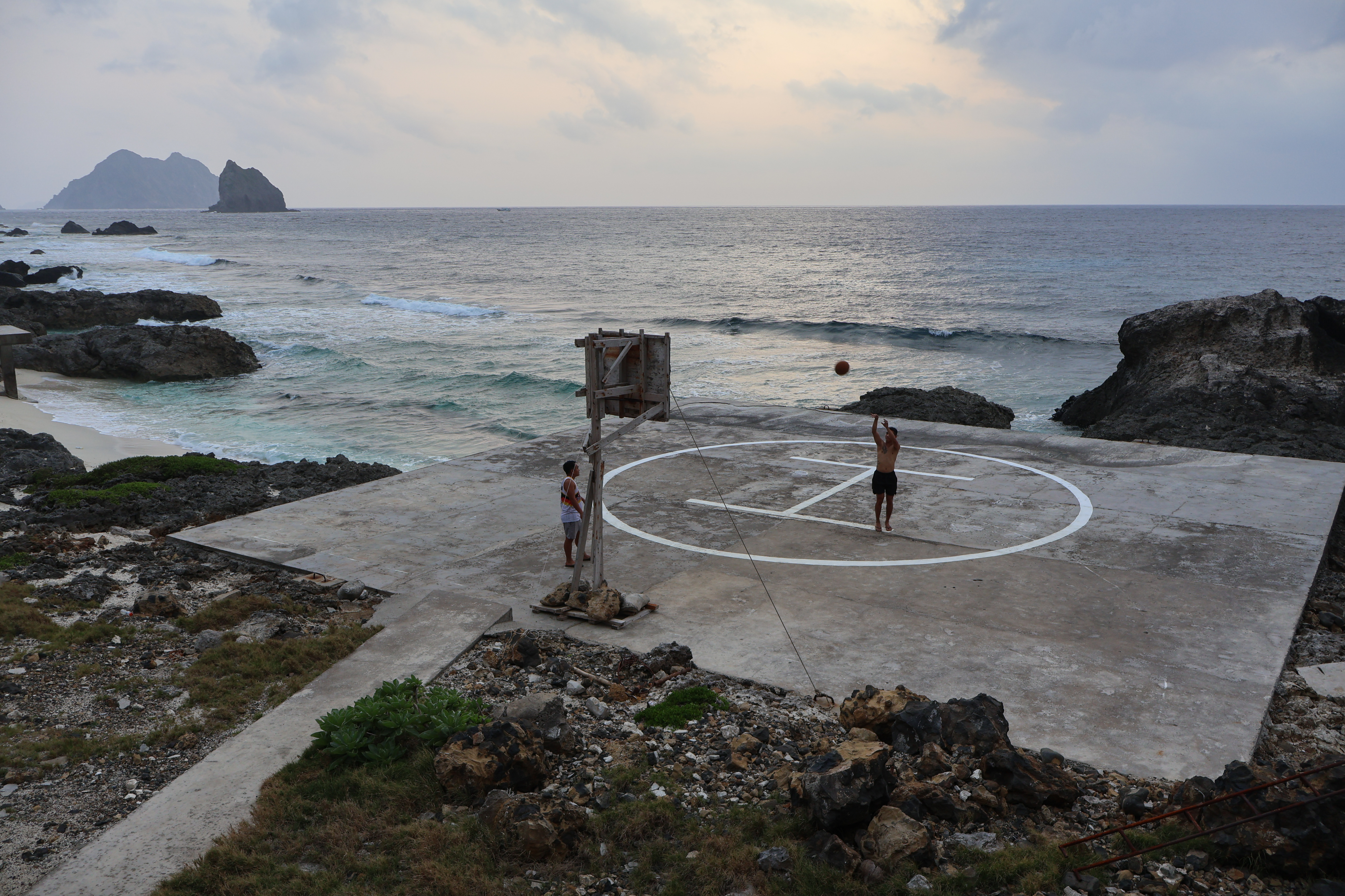 Two soldiers playing basketball on the Mavulis helipad. One is shooting at the basket. The sea is behind. And looks calm. 