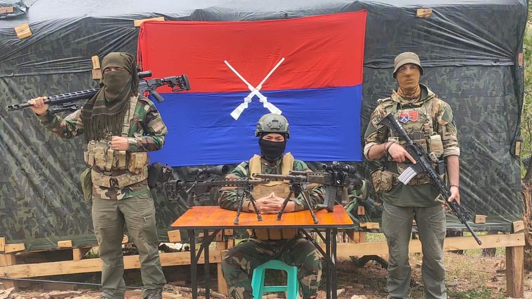 Azad, a US national, (left) and an unnamed British fighter (right) with the PDF Zoland. The two men are standing either side of a desk where another fighter is sitting with weapons on display. There is a flag behind them. The men are all masked.