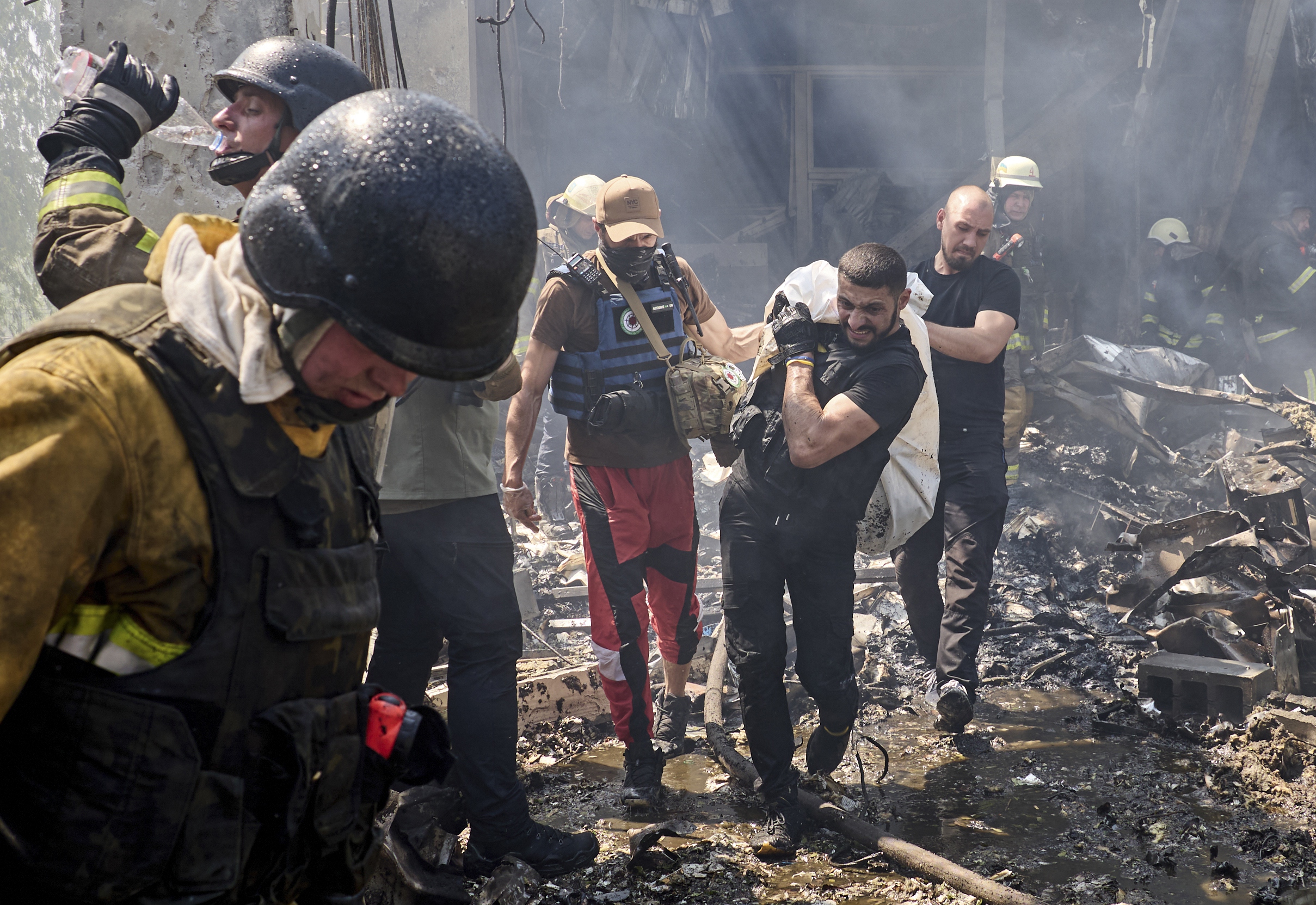 Search and rescue teams carry a victim from the smoking rubble of a Kharkiv printing factory hit in a Russian attack. Smoke is still rising from the debris. Firefighters are also at the site.