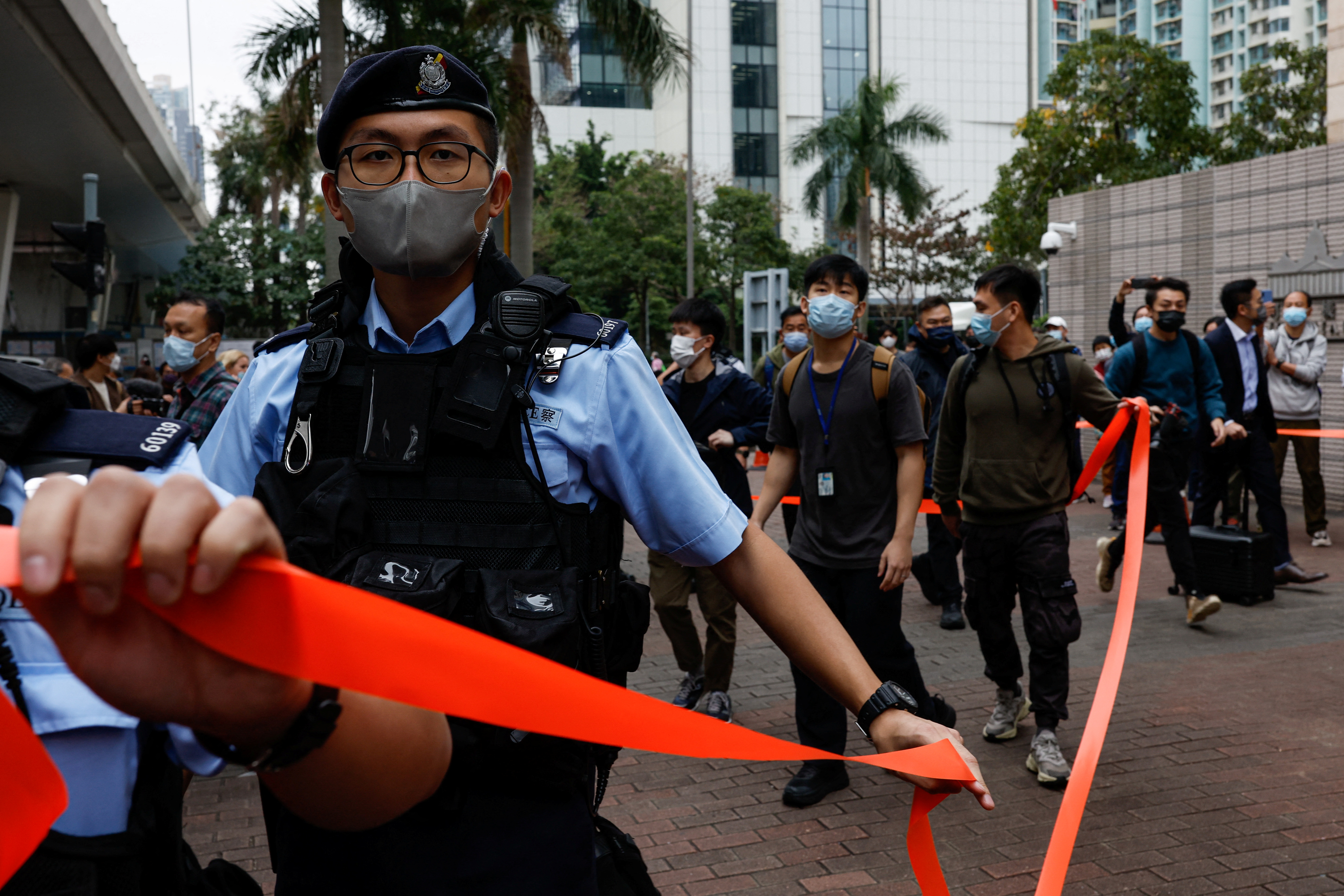 A police officer cordoning an area with red tape during one of the court hearings for the 47. There is a large group of supporters behind.