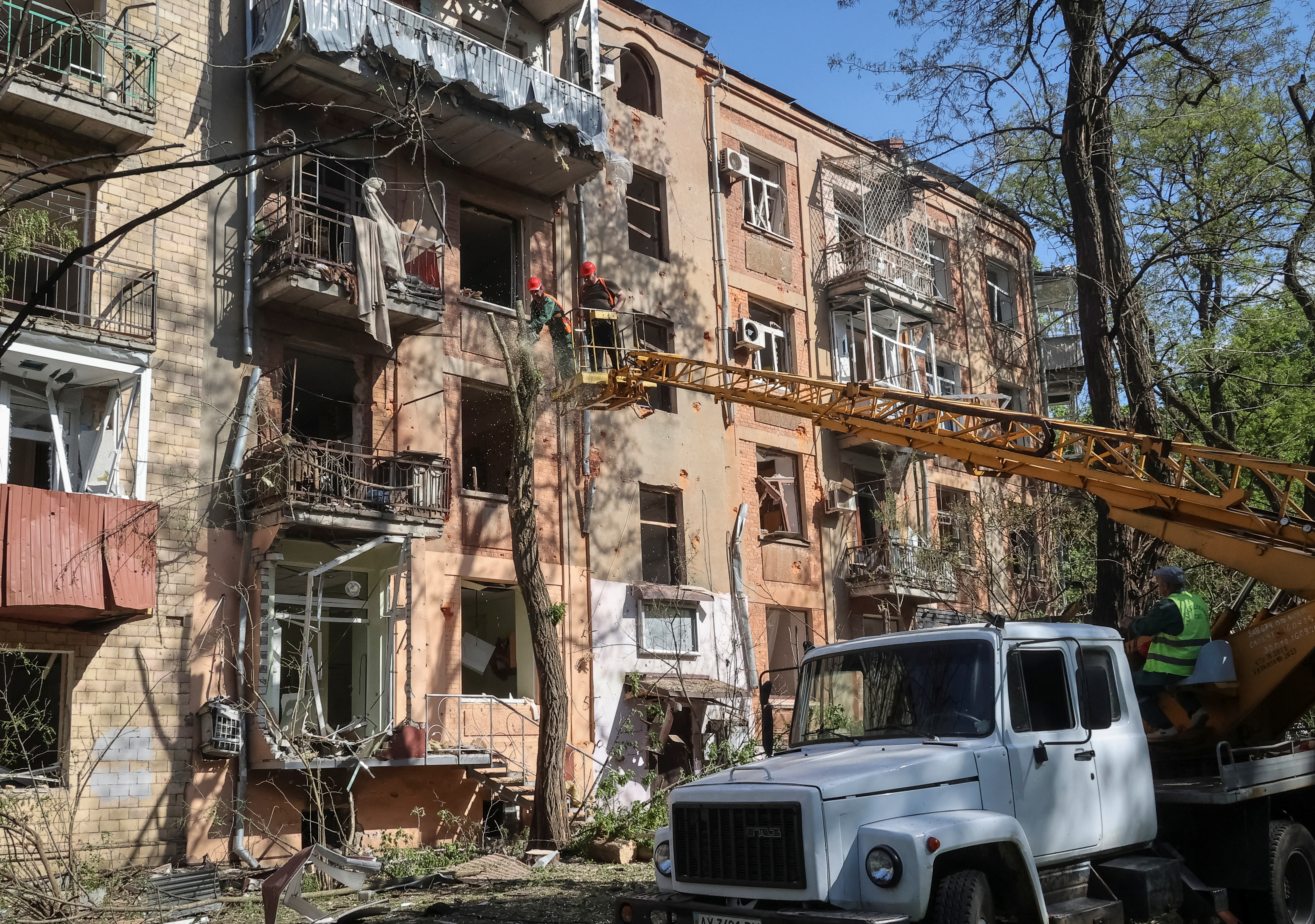 Emergency workers at an apartment building in Kharkiv that was hit in a Russian attack. The building is four storeys high and painted a peachy pink. The windows have been blown out. The workers are on a crane attached to a white truck parked in front.