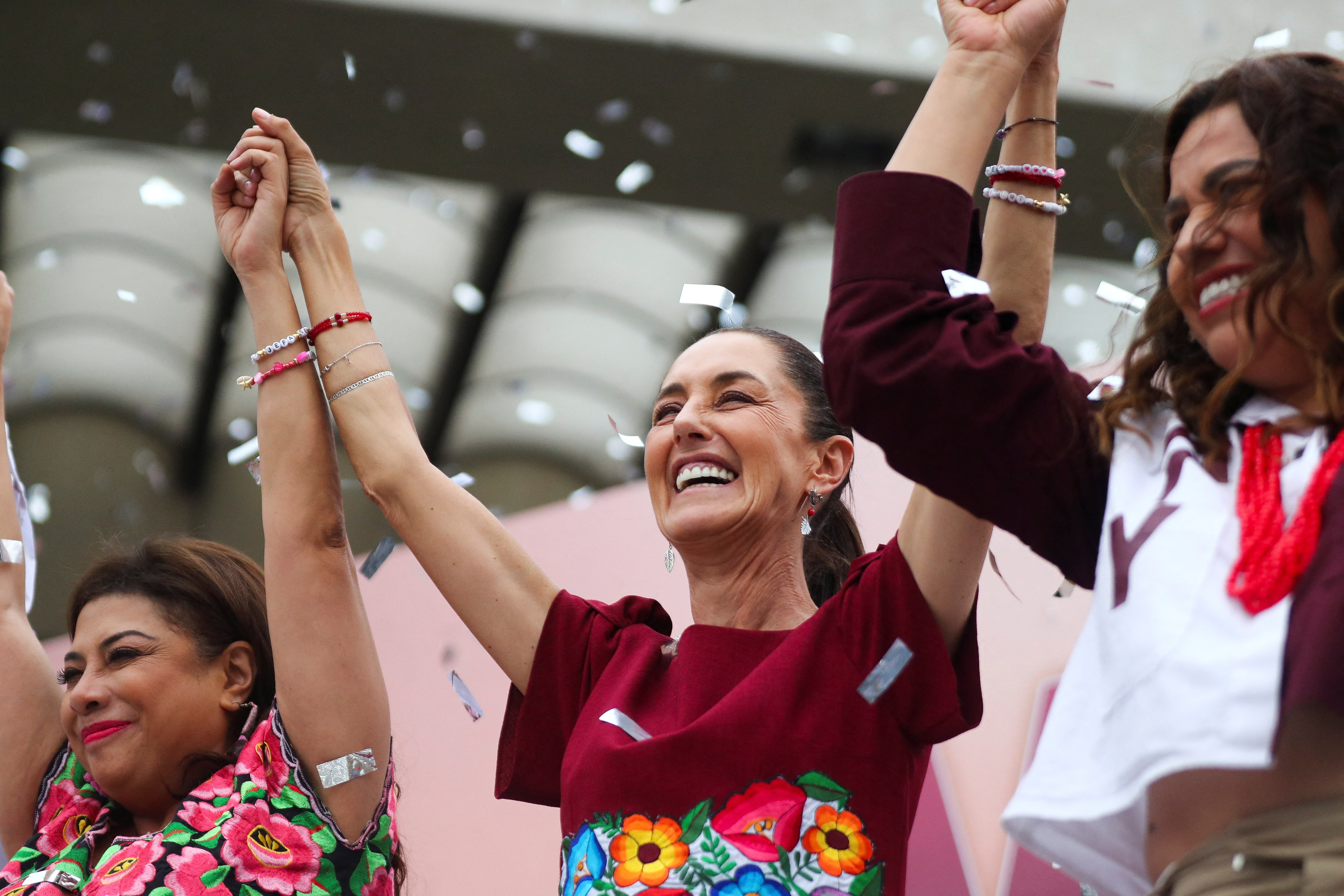 Claudia Sheinbaum lifts her arms and holds hands with two other women on stage at a campaign rally, as confetti falls.