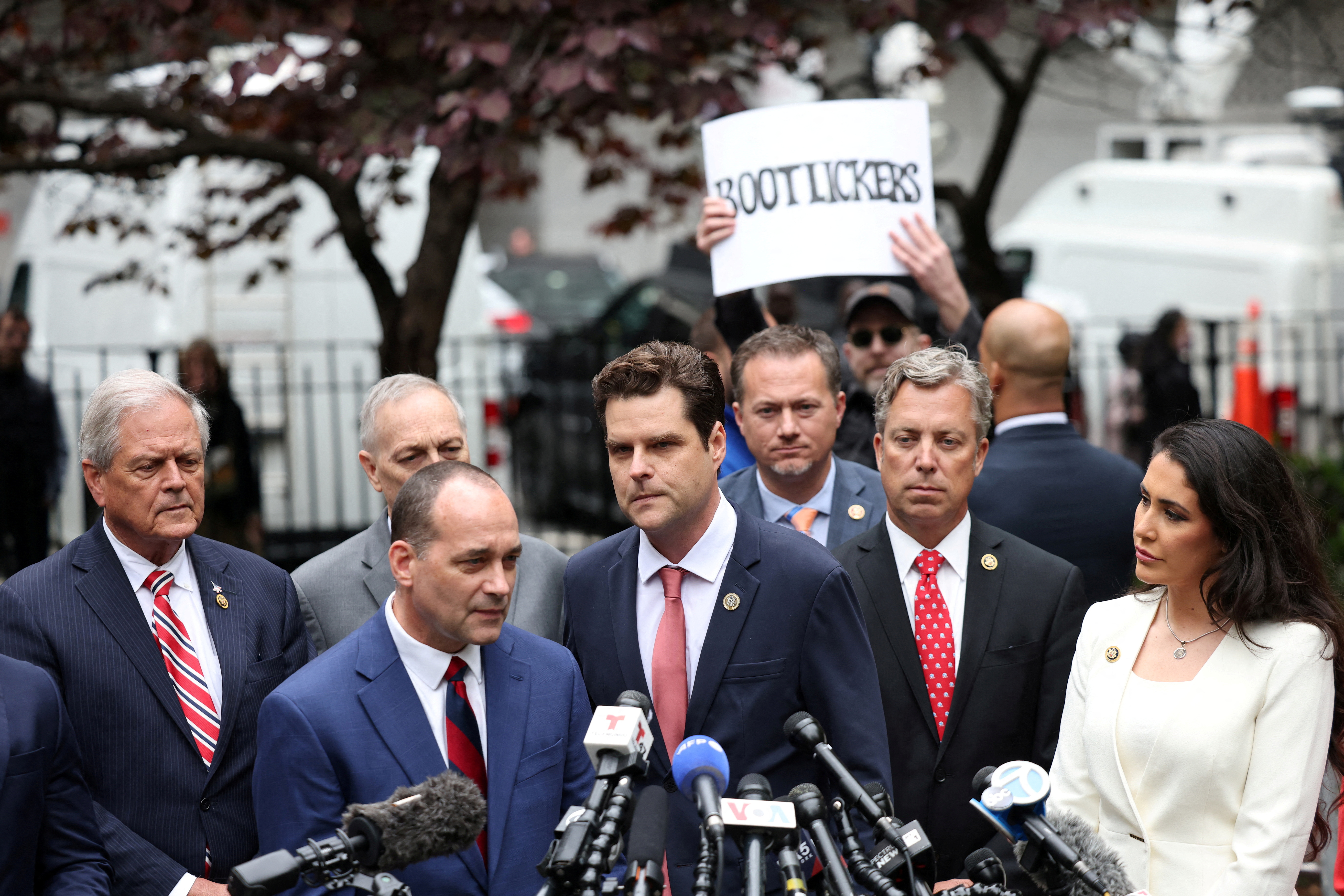 Matt Gaetz speaks to reporters outside the Manhattan Criminal Court, surrounded by reporters. Behind him, a protester holds up a sign that reads, "Bootlickers."