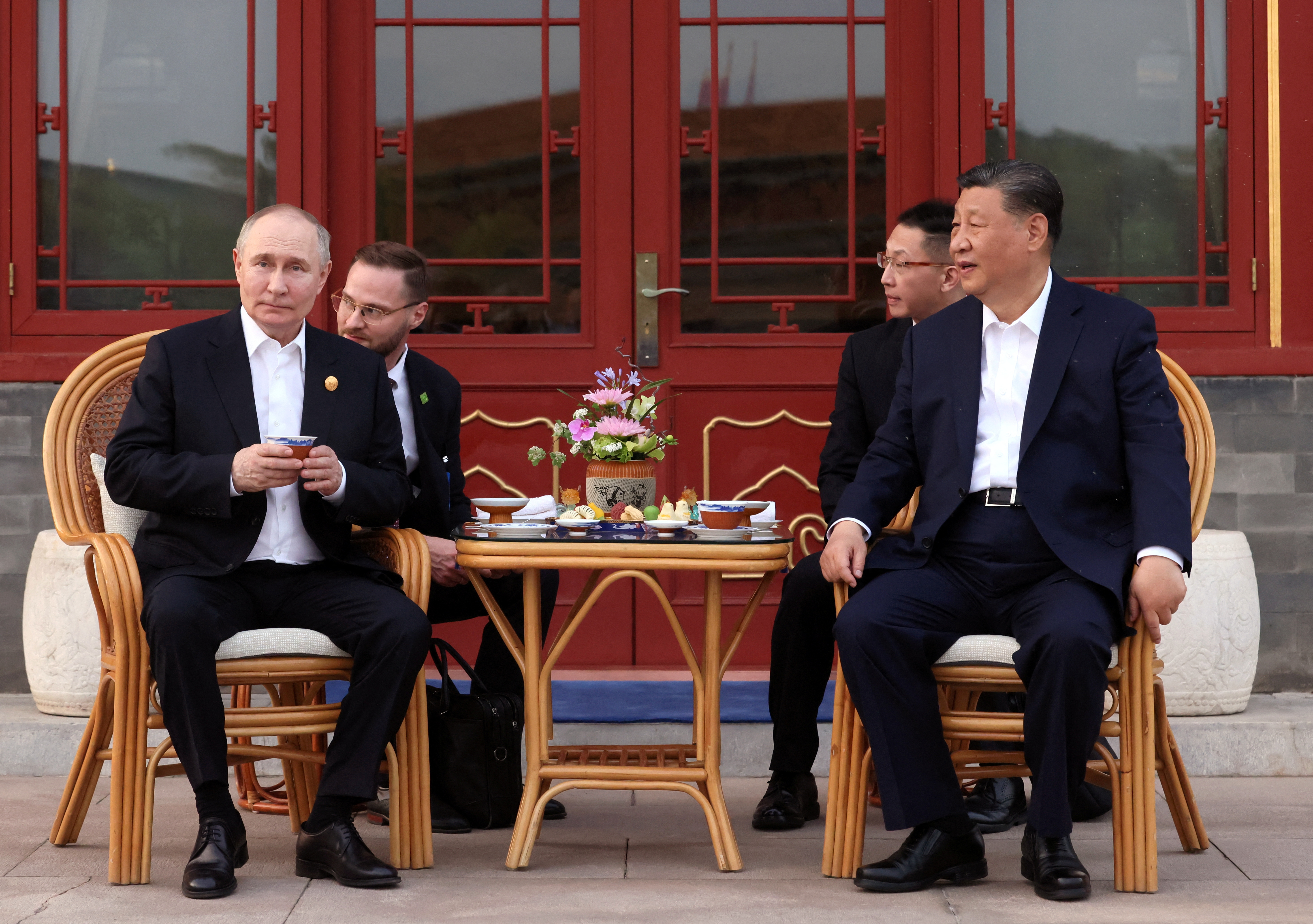 Vladimir Putin and Xi Jinping having tea outside in a Beijing park. They are sitting on a verandah on wicker chairs with a table between them. Their intepreters are behind them. Putin is holding his tea cup.