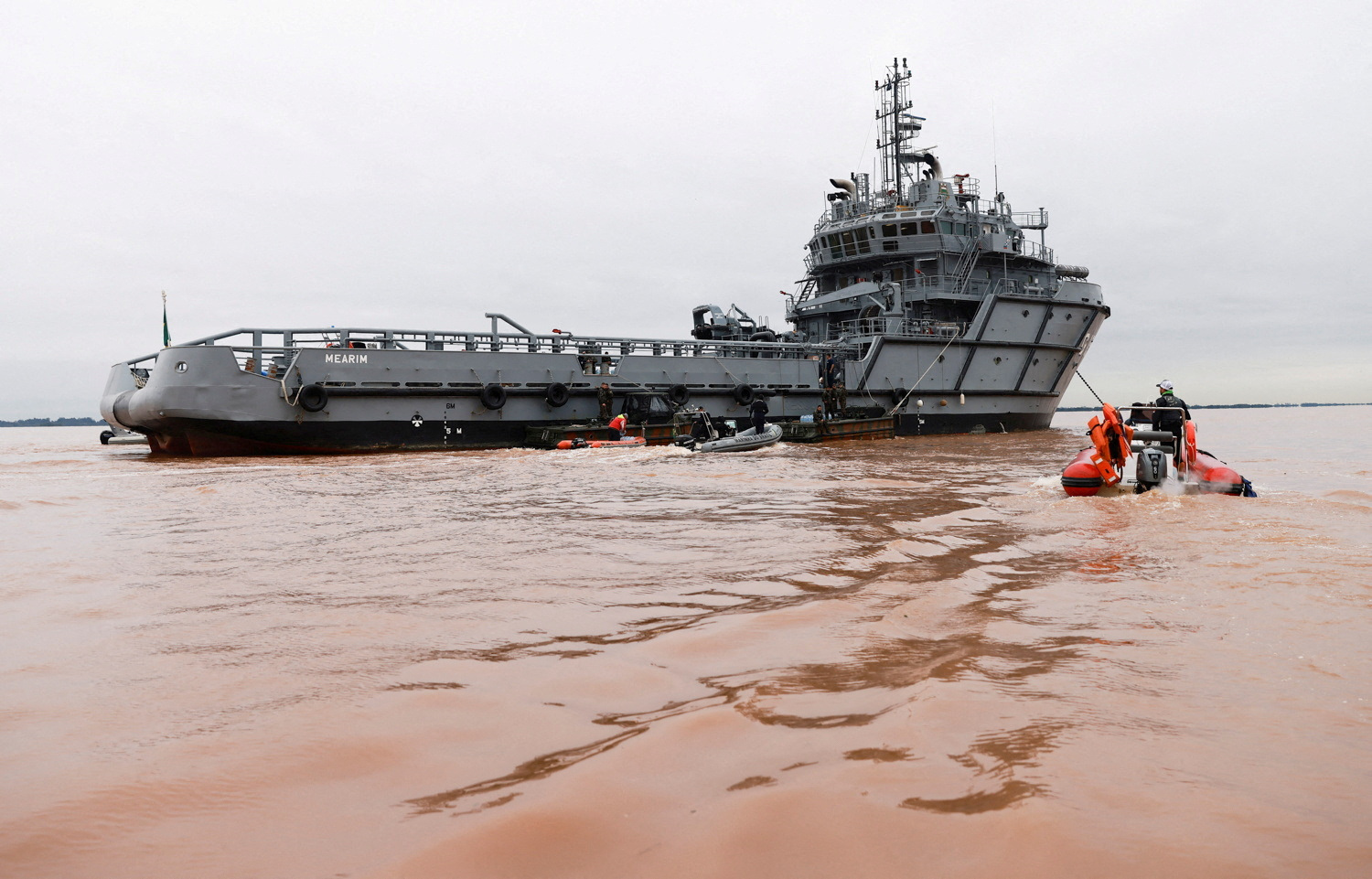 A military ship sits in southern Brazil's muddy floodwaters, with smaller vessels coming to and from it.