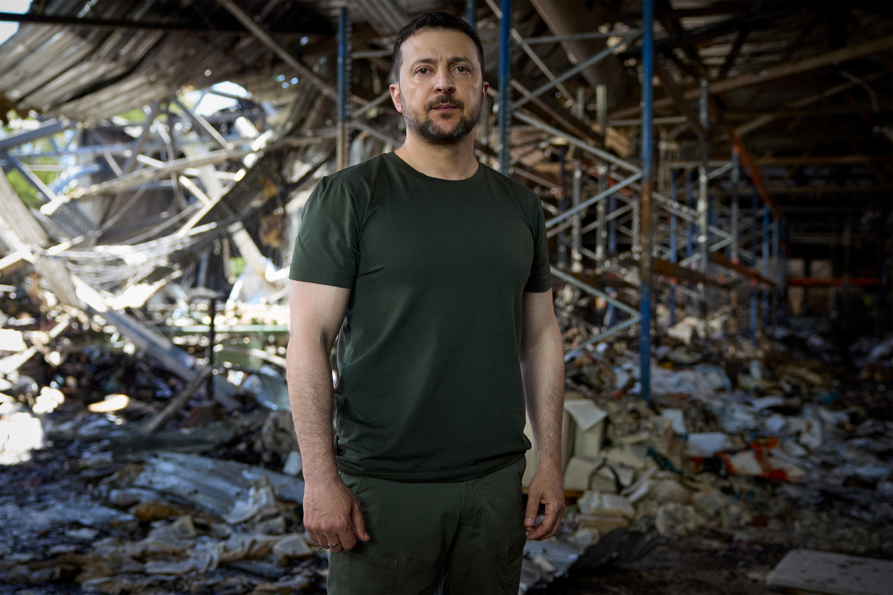 Ukrainian President Volodomyr Zelenskyy standing inside the ruins of a printing works attacked by Russia. He is wearing a khaki t-shirt and dark trousers. The building is charred by fire with debris lying on the floor. The roof behind him has collapsed.