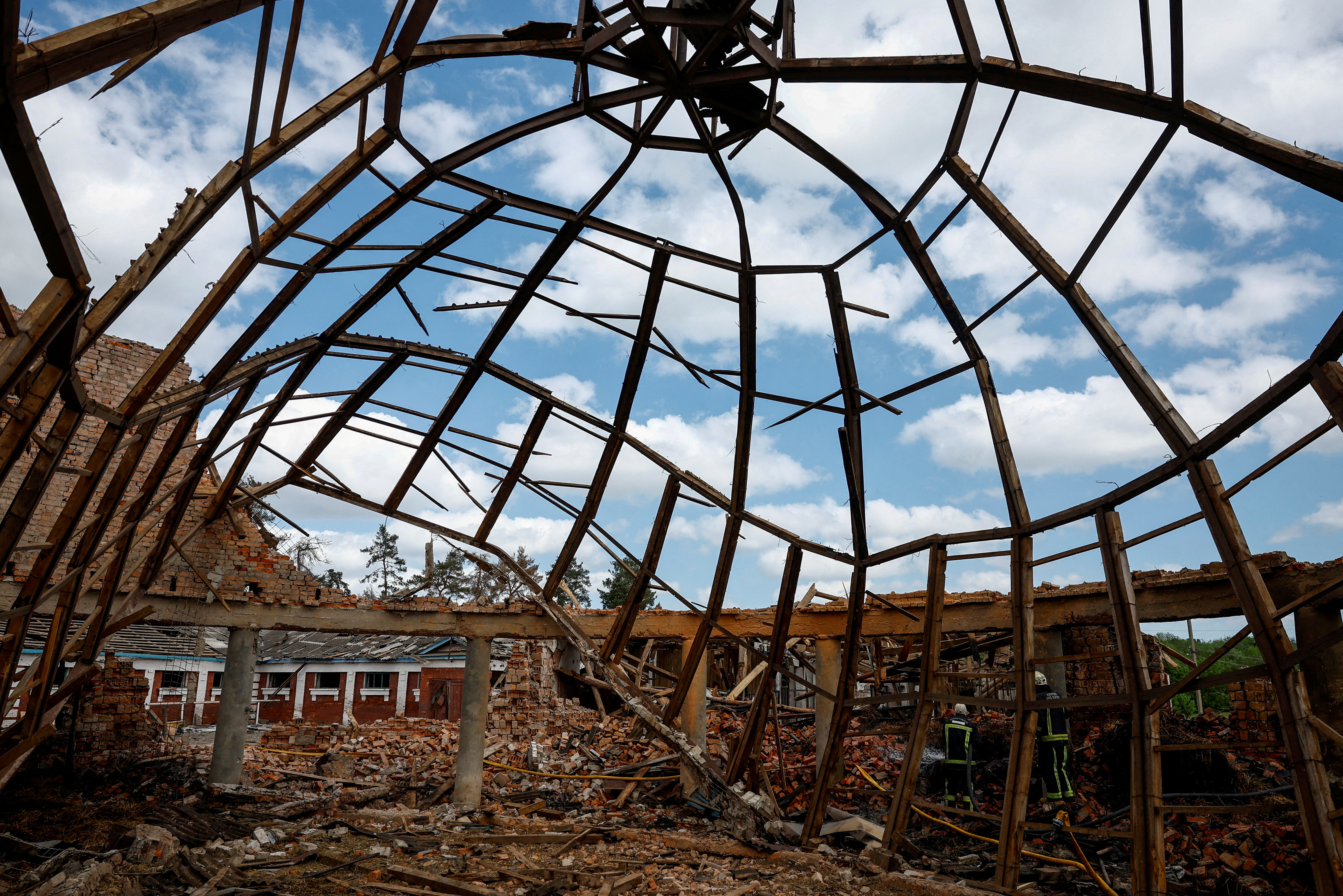 A damaged building at a riding school after a Russian attack outside Kharkiv. The roof is mangled. There is rubble all over the ground. Firefighters are working at the site.
