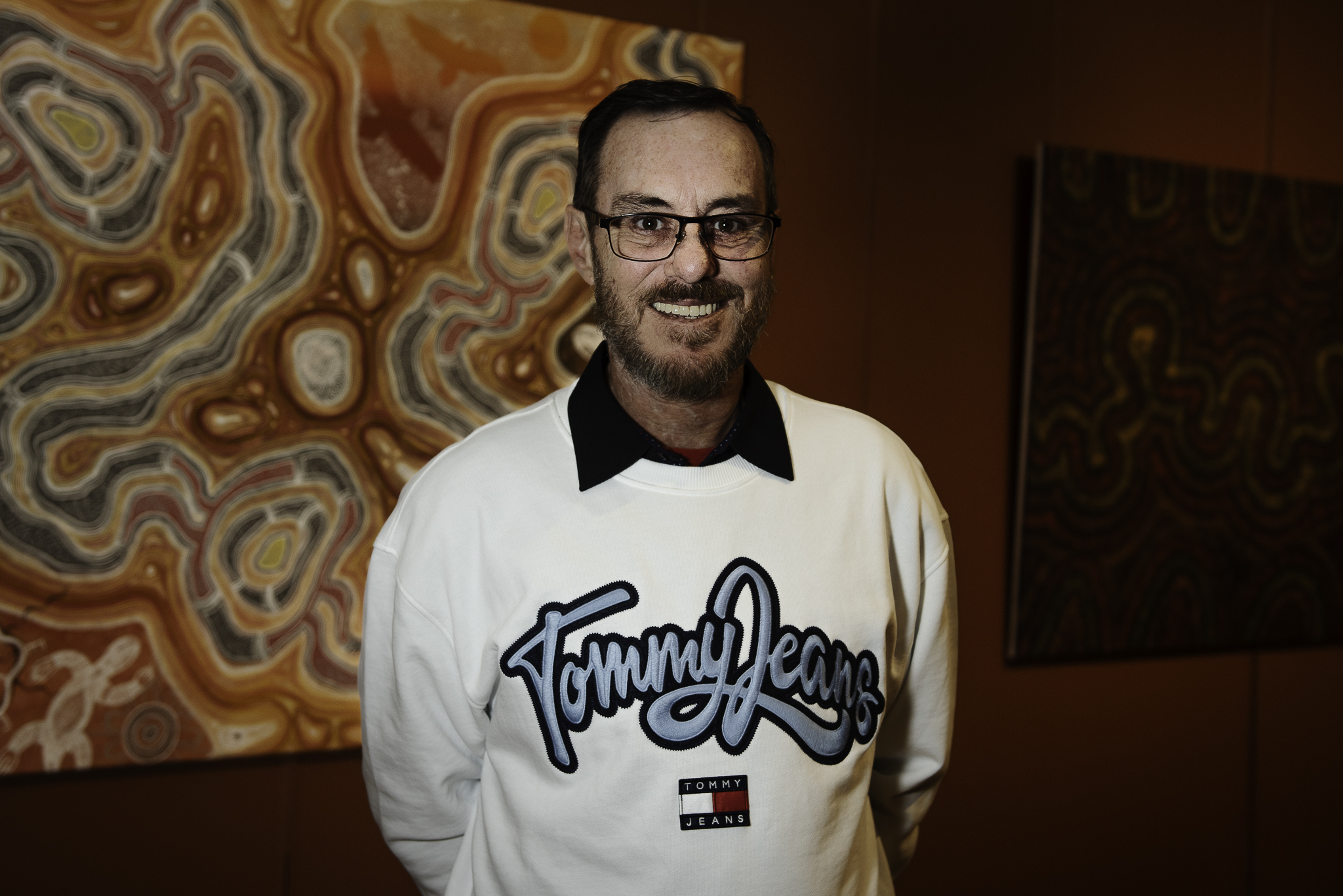 Ash Thomas. He is wearing a black shirt with a white logo sweatshirt. He has dark hair and glasses. He's standing in front of his art