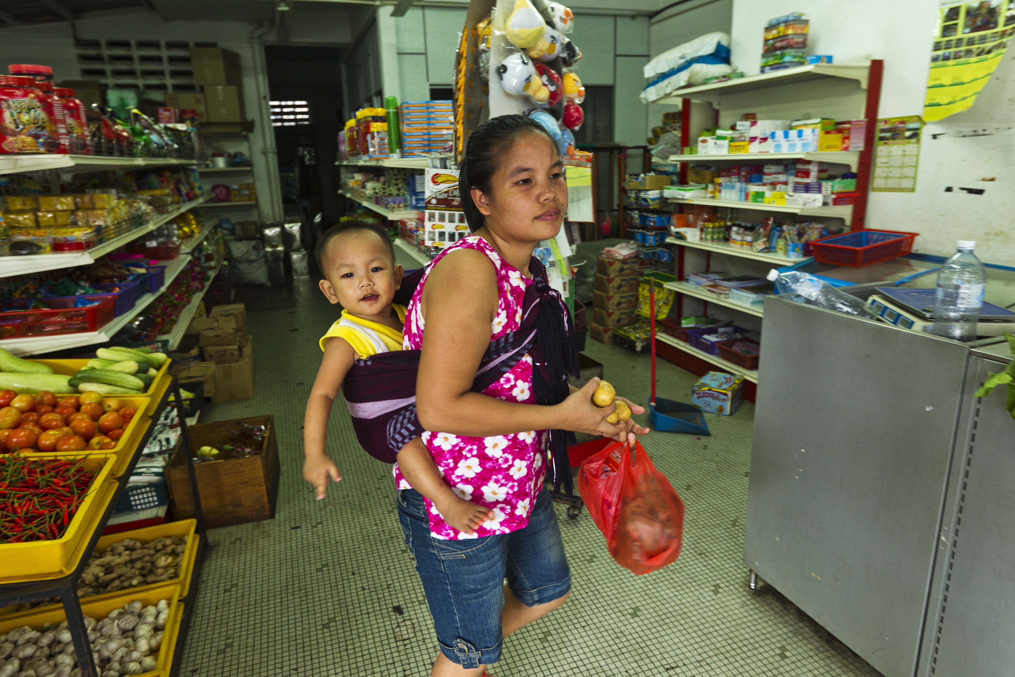 An ethnic Chin woman from Myanmar inside a sundry shop in Malaysia. She has her young child strapped to her back in a sarong. She is carrying a small plastic bag.